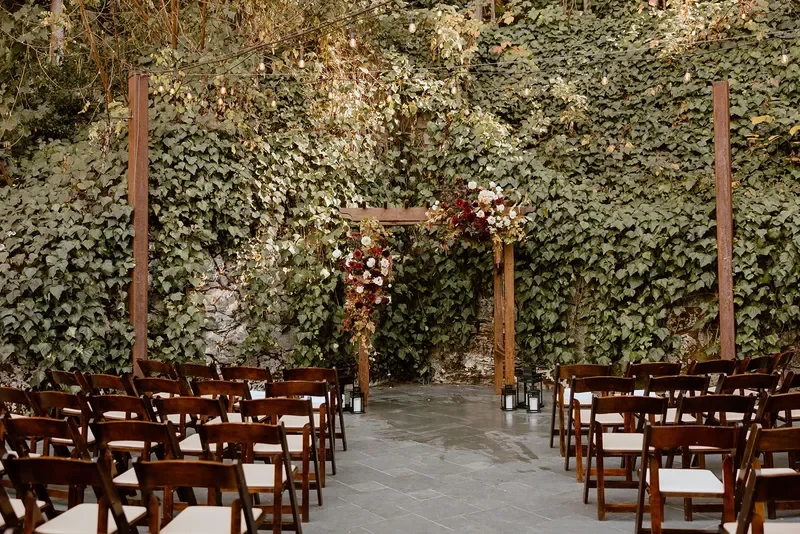 Outdoor wedding ceremony setup with wooden chairs arranged facing a floral arch against a stone wall covered in ivy.