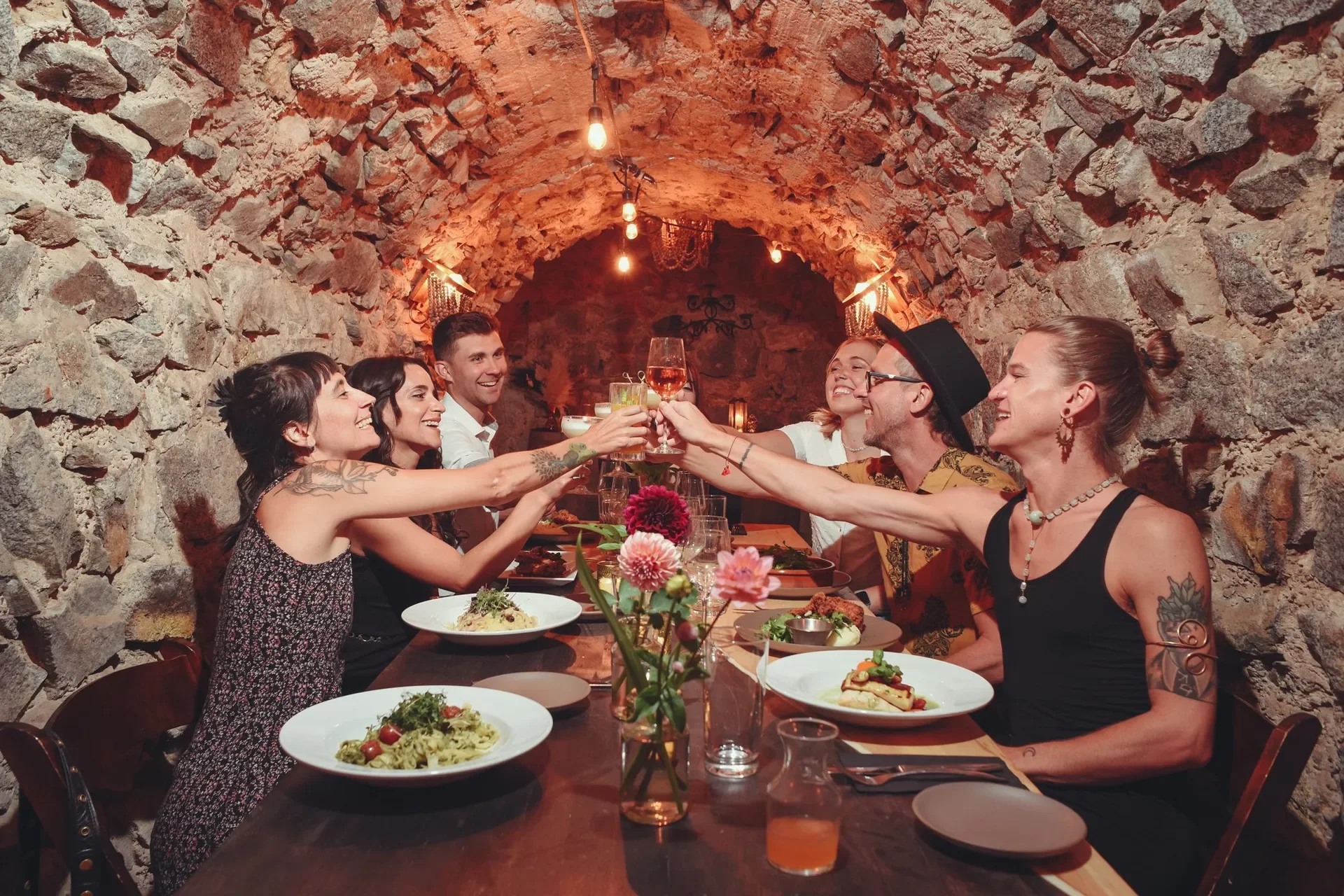 A group of friends enjoying dinner together in a rustic stone-walled restaurant, raising their glasses in a toast.