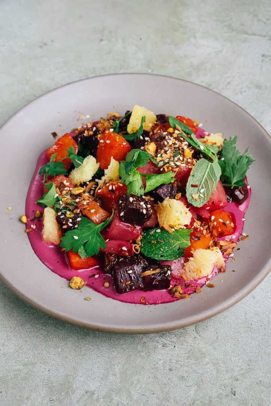 A colorful beet salad with cherry tomatoes, pineapple chunks, fresh herbs, and crushed nuts on a pink sauce base, served on a gray plate.