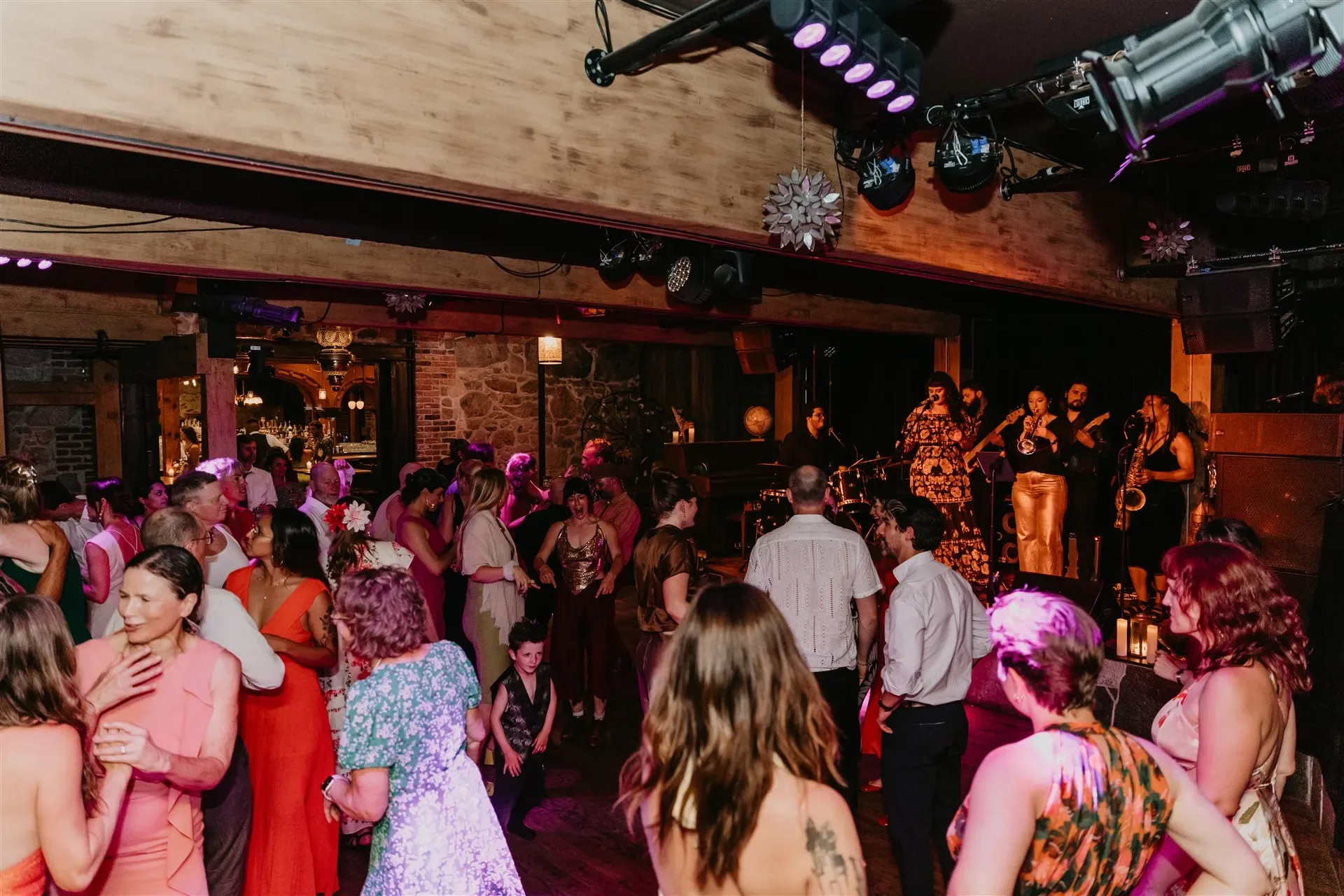 Crowd of people dancing and socializing at a lively indoor event with a band performing on stage in the background, decorated with warm lighting and floral accents.
