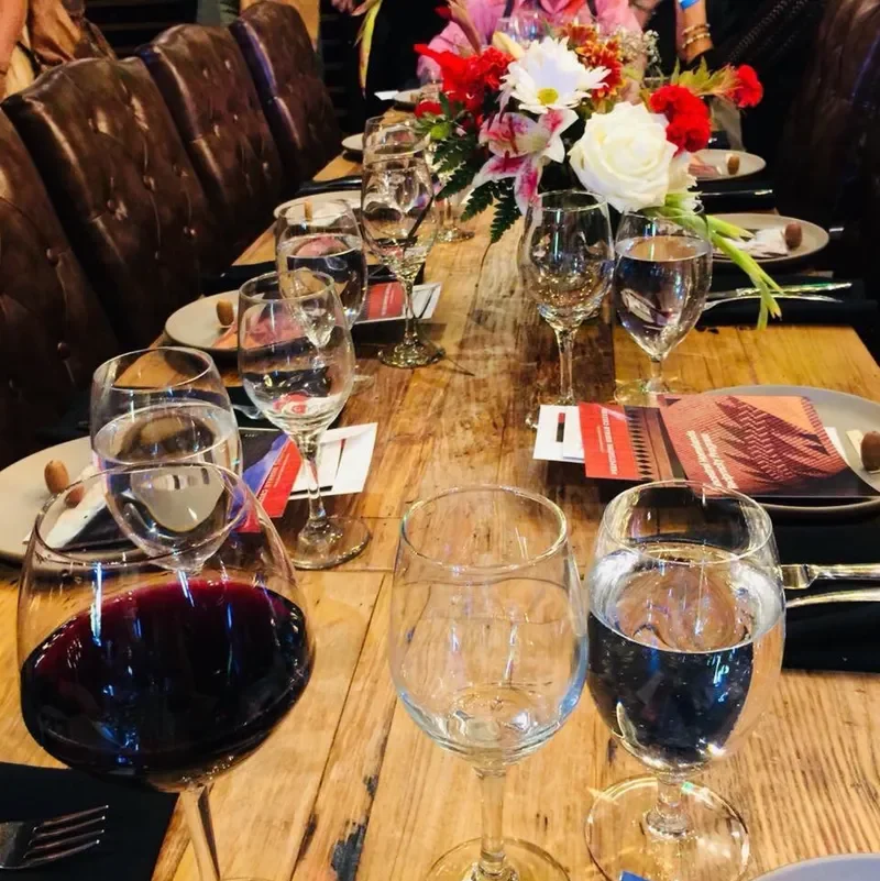 A long wooden banquet table set for a formal dining event with glasses of water, wine, and wine glasses, floral centerpiece with pink, white, and red flowers, plates with bread rolls, and party favors, with brown leather chairs along the sides.