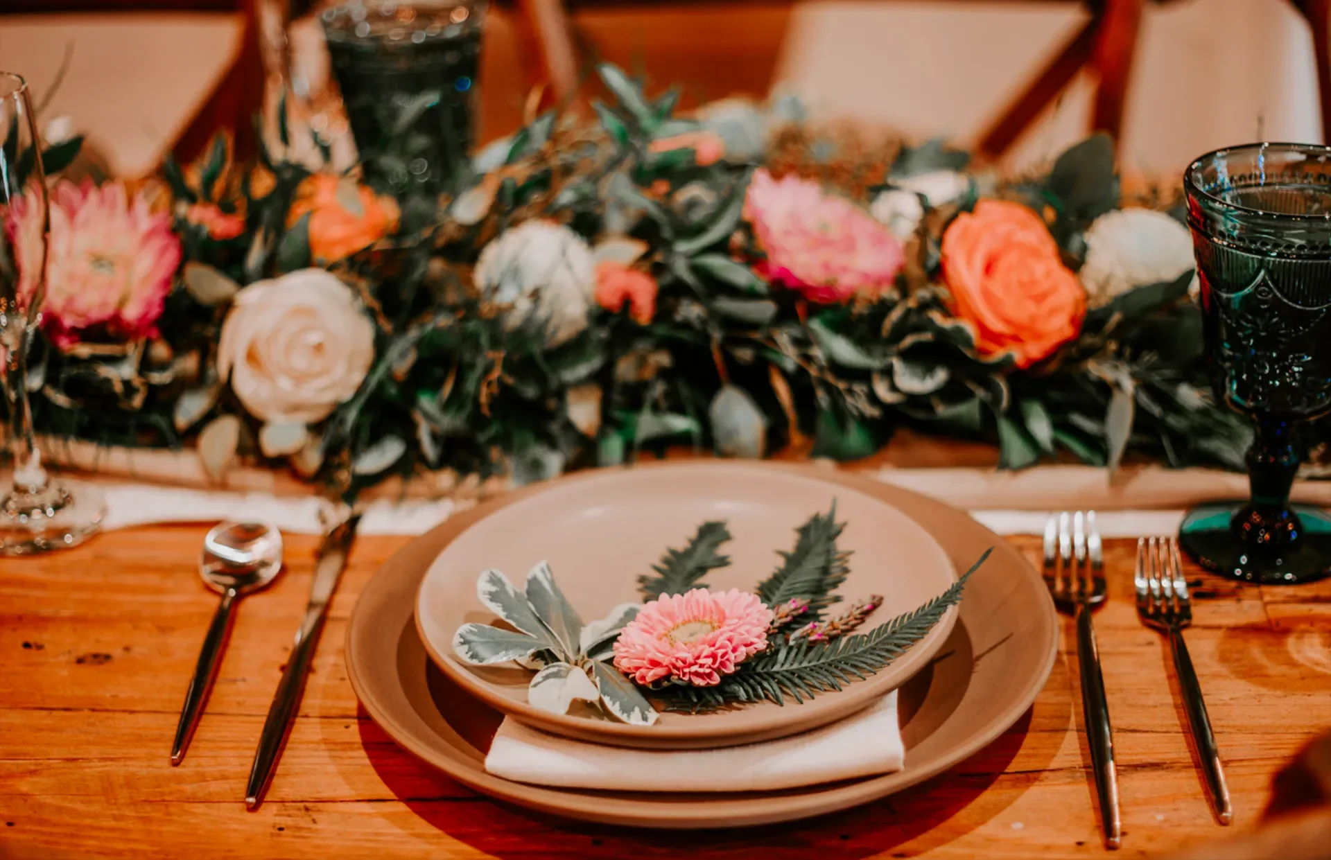 Table setting with a beige plate and a floral arrangement on top, surrounded by silverware and black glasses, with a floral centerpiece in the background.