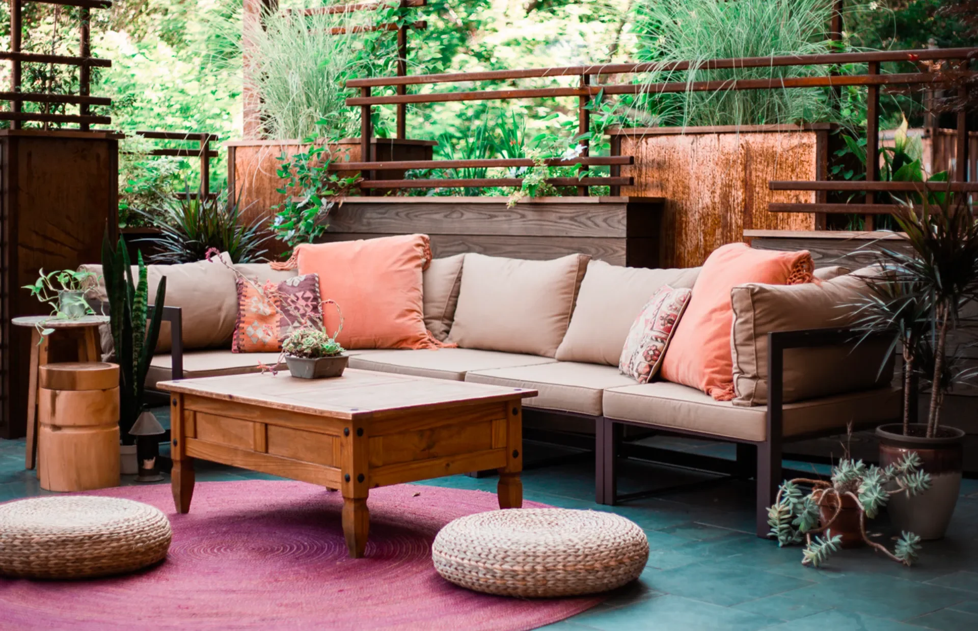 Outdoor patio with a beige L-shaped sofa, orange and patterned throw pillows, a wooden coffee table, woven poufs, and potted plants. Wooden railing and lush green foliage in the background.