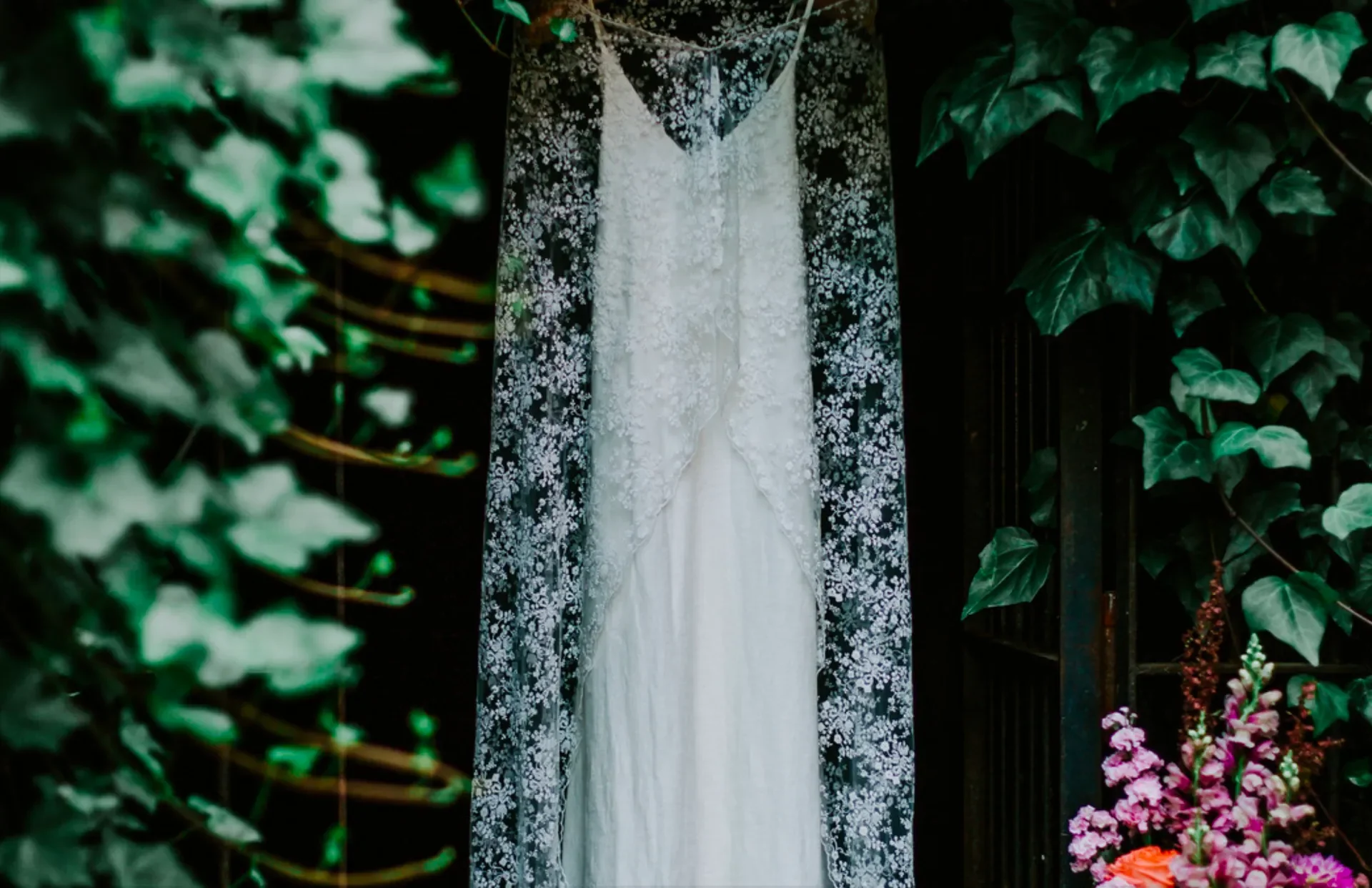 A white wedding gown with lace details hanging on a black background with green foliage and pink flowers on the sides.