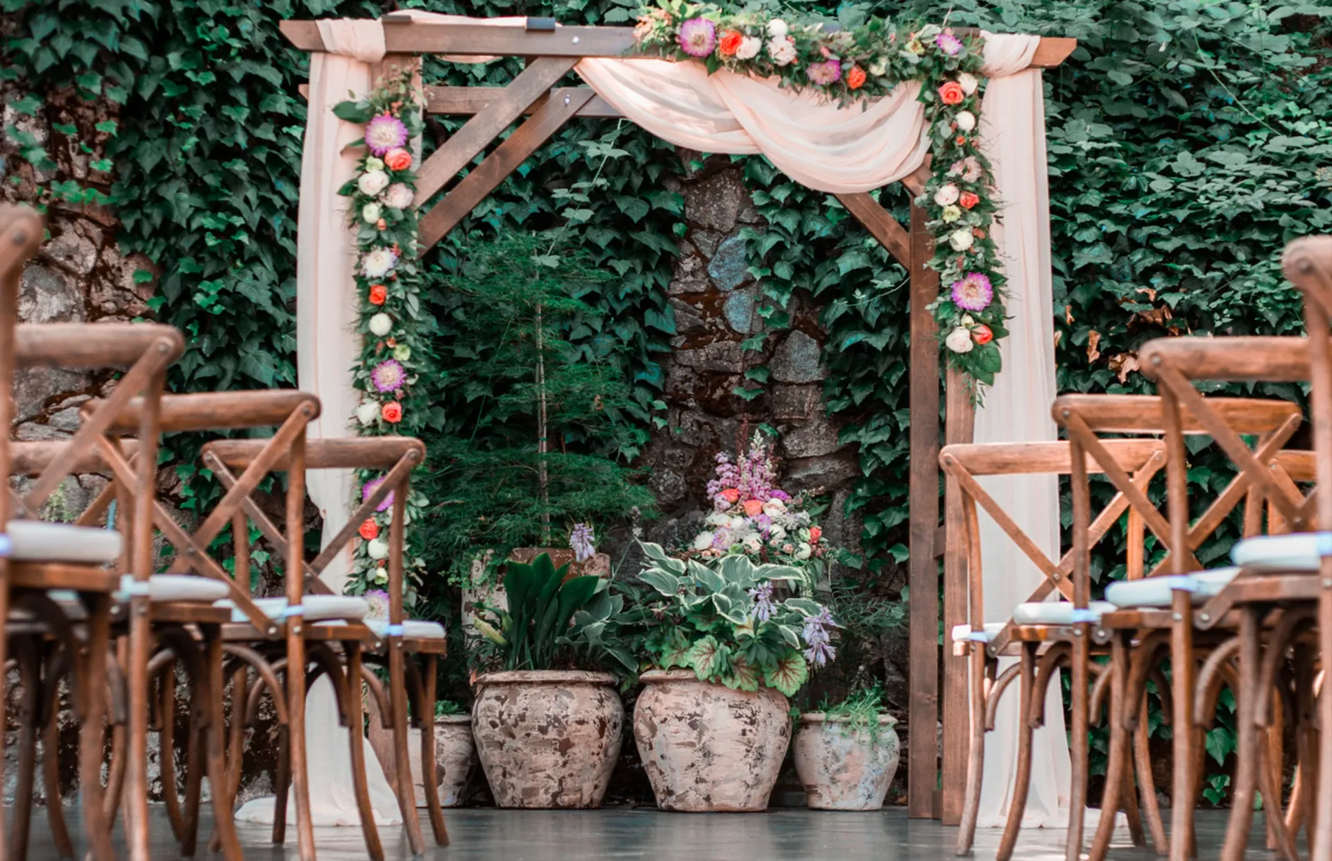A decorated wedding arch with pink and white flowers and draped fabric, set against a backdrop of ivy-covered stone wall, with potted plants at its base and wooden chairs arranged for a ceremony.