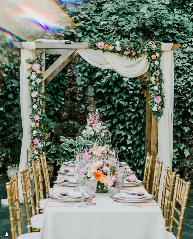 A decorated outdoor dining table set for a wedding or special event, with floral centerpieces, gold chairs, and an arch draped with fabric and flowers, surrounded by green ivy.