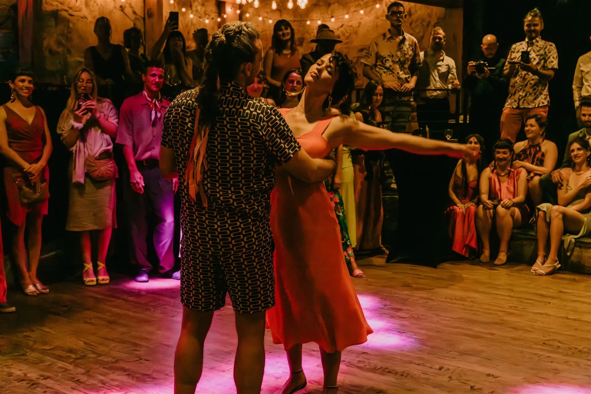 A dance couple is performing in front of an audience in a lively, dimly lit venue with warm lighting and wooden floors. The woman in an orange dress is extending her arm, while the man in a patterned shirt and shorts faces her, her head tilted back s