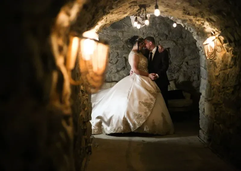A bride and groom sharing a kiss in a stone-walled basement or cellar, decorated with hanging lights.