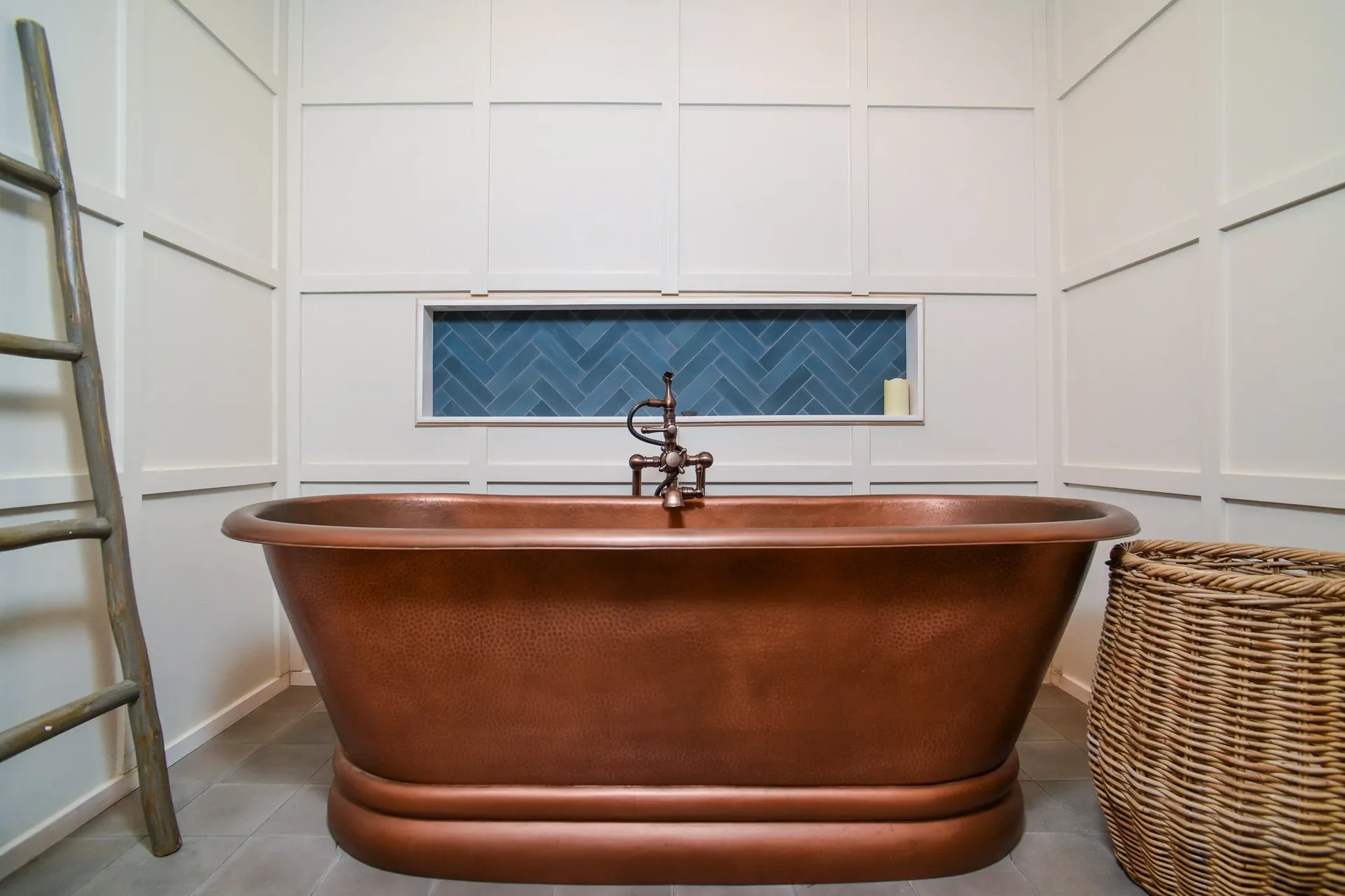 Copper bathtub in a bathroom with white paneled walls, a blue tile window, a ladder on the left, and a woven basket on the right.