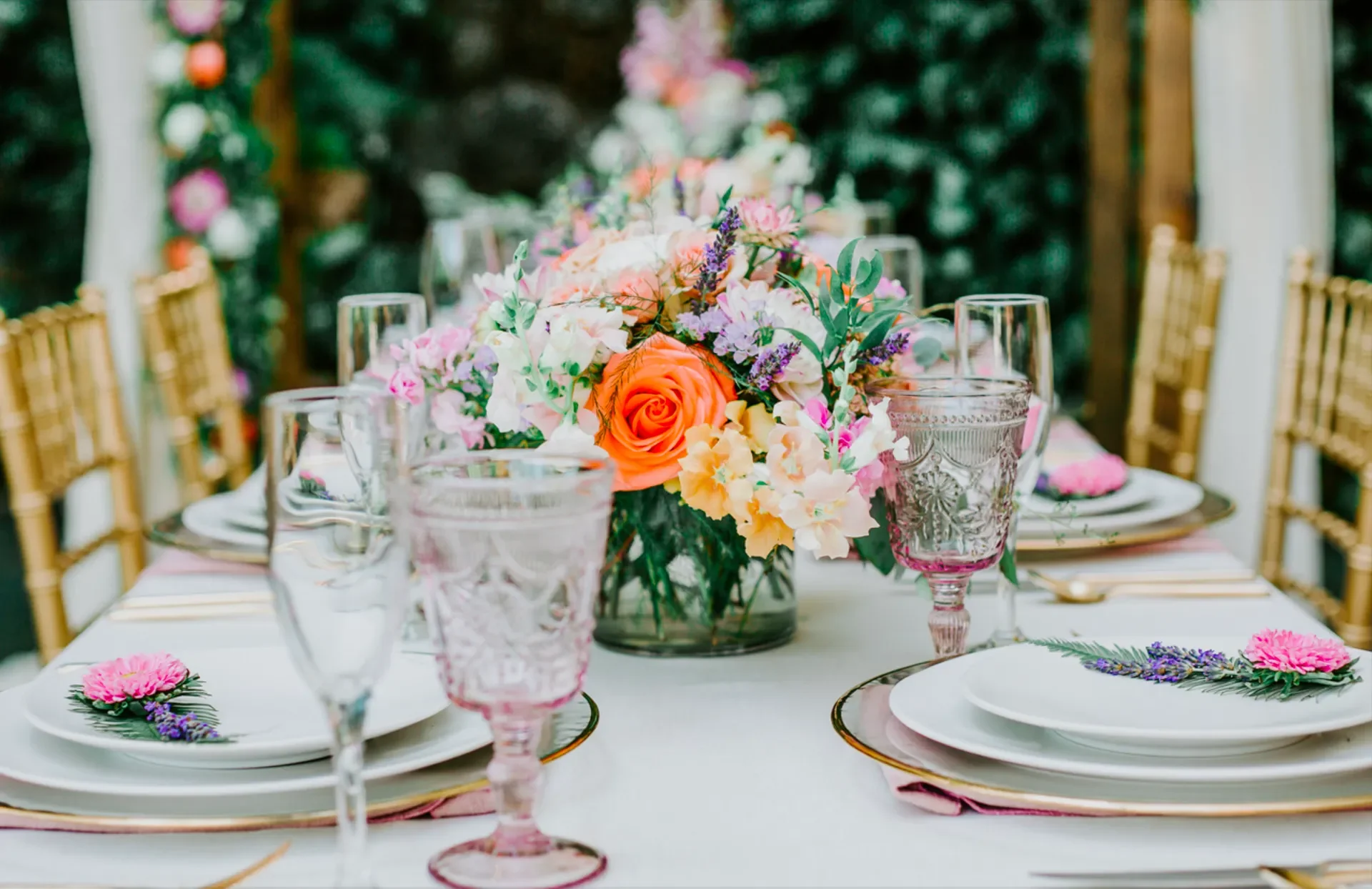 Elegant table setting with a bouquet of pink, orange, and purple flowers in a glass vase at the center, surrounded by gold chairs, white plates with floral napkins, pink and clear drinking glasses, and dessert forks, set outdoors with a blurred green