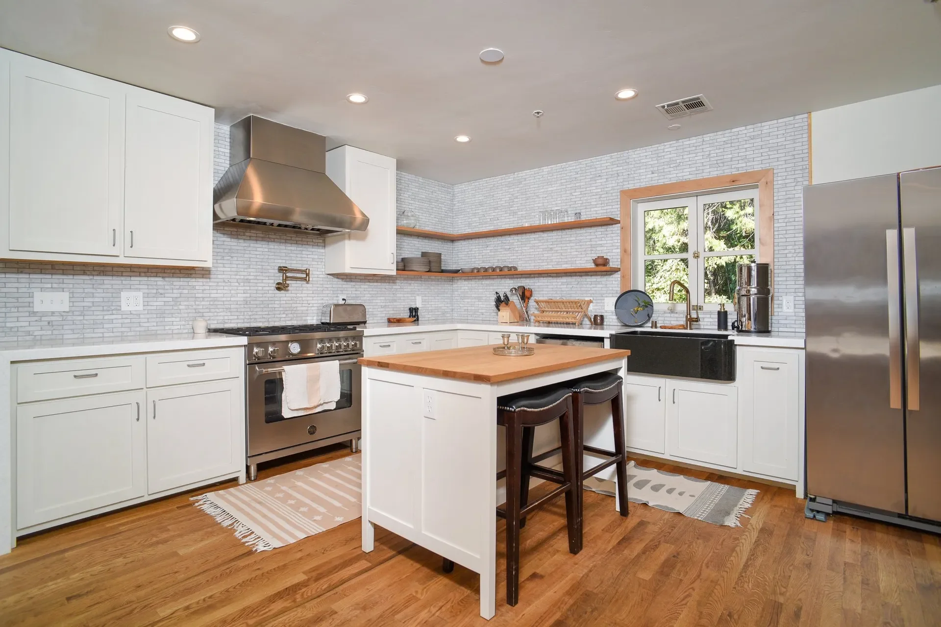 A modern kitchen with white cabinets, a wooden kitchen island, stainless steel appliances, open wooden shelves, a black farmhouse sink, hardwood floors, and a window overlooking greenery.