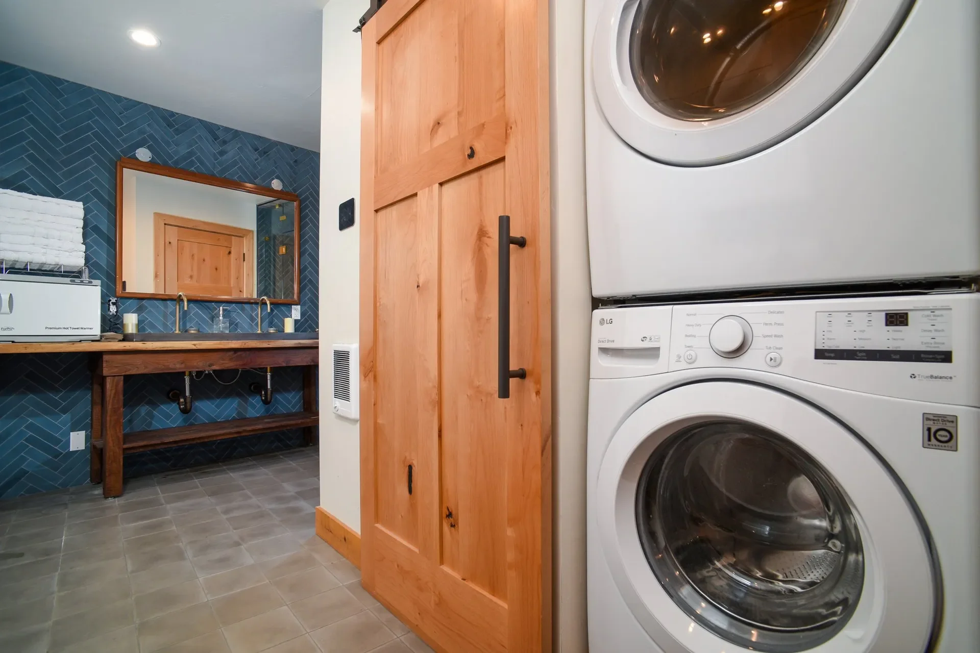 A laundry room with front-loading washer and dryer, a wooden cabinet with black handles, a wooden framed mirror above a wood countertop with double sink, a blue tiled wall, and a gray tiled floor.
