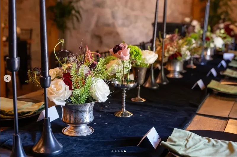 A decorated dining table with black tablecloth, multiple silver flower arrangements, black candles in holders, gold-rimmed plates, and folded napkins, set for a formal event.