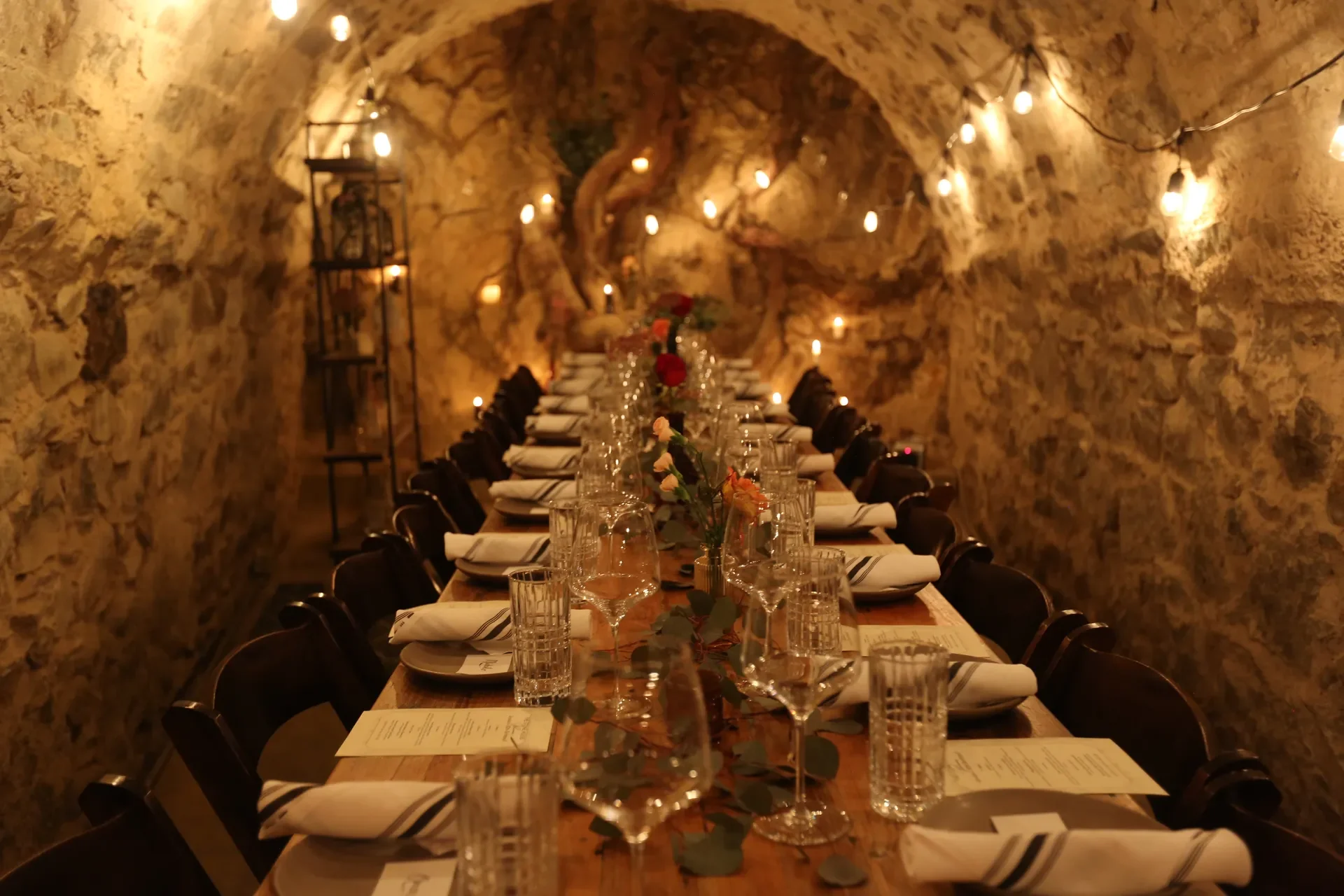 Long dining table set for a meal inside a stone-walled cellar with string lights overhead and centerpieces of flowers and greenery