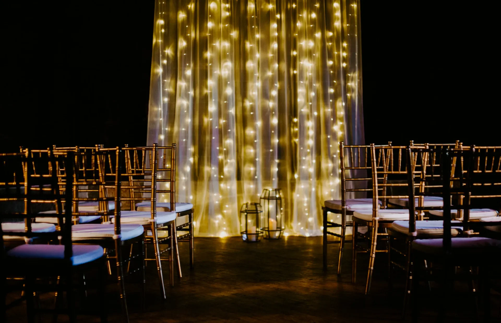 An indoor event space set up with rows of gold Chiavari chairs facing a backdrop of soft yellow and white fairy lights on dark curtains, with decorative lanterns on the floor in front of the backdrop.