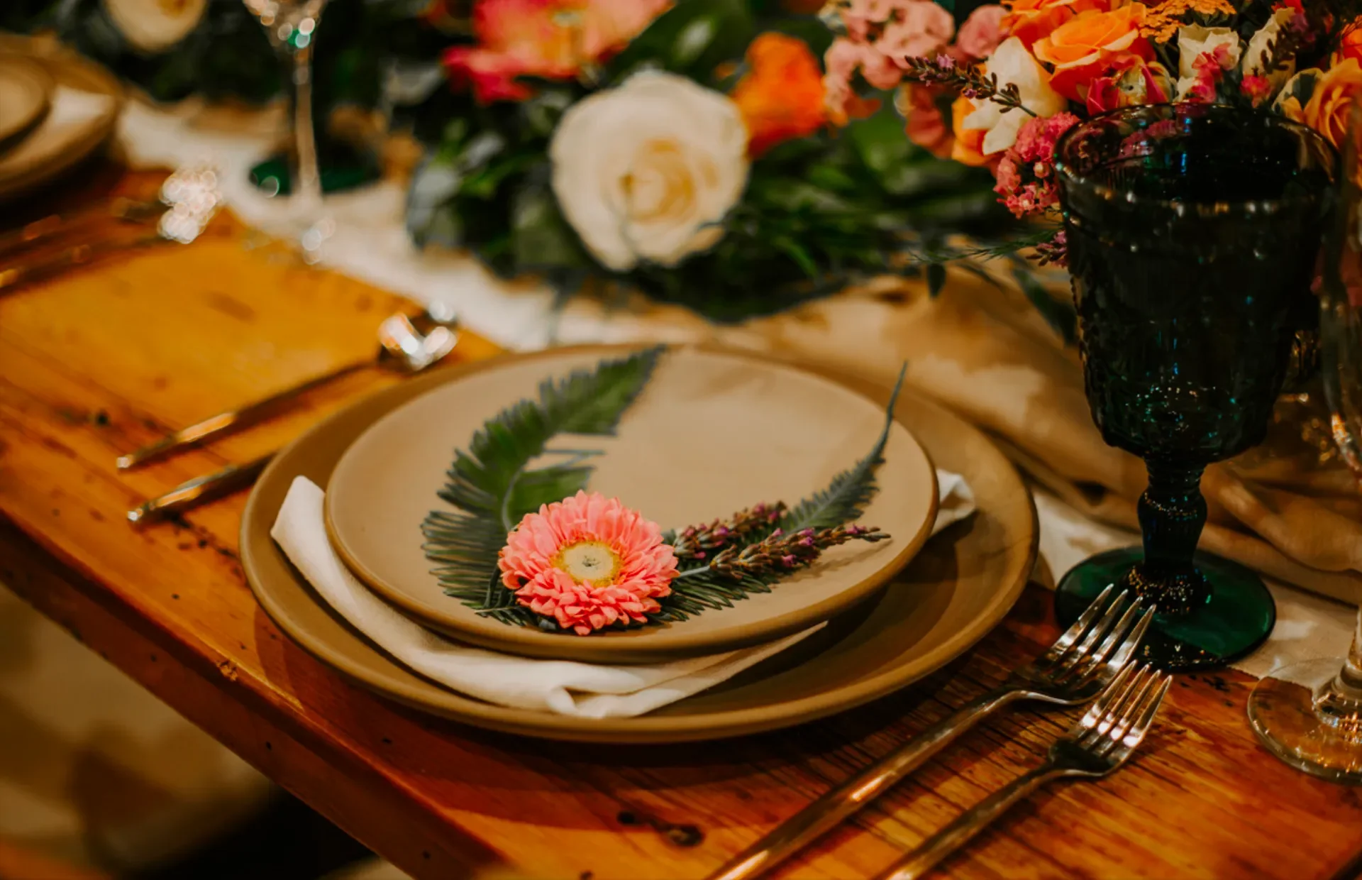A table setting with beige plates, a white cloth napkin, silver cutlery, a dark green goblet, and a floral arrangement with pink and white flowers and greenery.