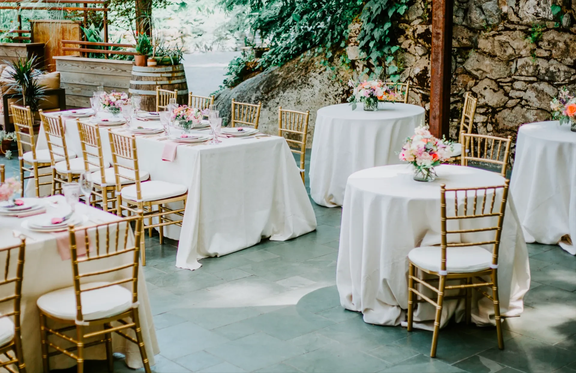 Elegant indoor dining area with round and rectangular tables covered in white tablecloths, decorated with pink floral centerpieces, surrounded by gold chairs with white cushions, set up for a formal event or wedding reception.