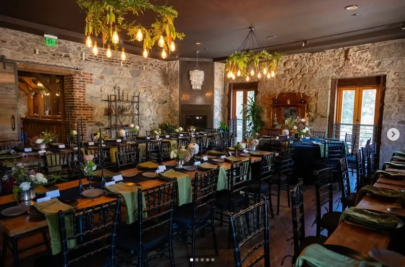 A rustic indoor dining area decorated for a special event with long tables set with plates, napkins, and floral centerpieces, surrounded by black wooden chairs, with hanging lights and greenery overhead, stone walls, and large windows.