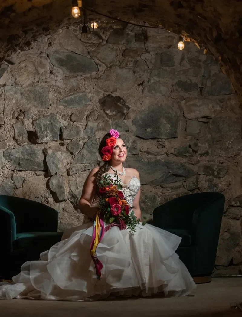 A bride in a white wedding gown sits on the floor in front of a rustic stone wall, holding a bouquet of vibrant pink, red, and orange flowers with colorful ribbons, smiling and looking upward, illuminated by warm hanging lights.