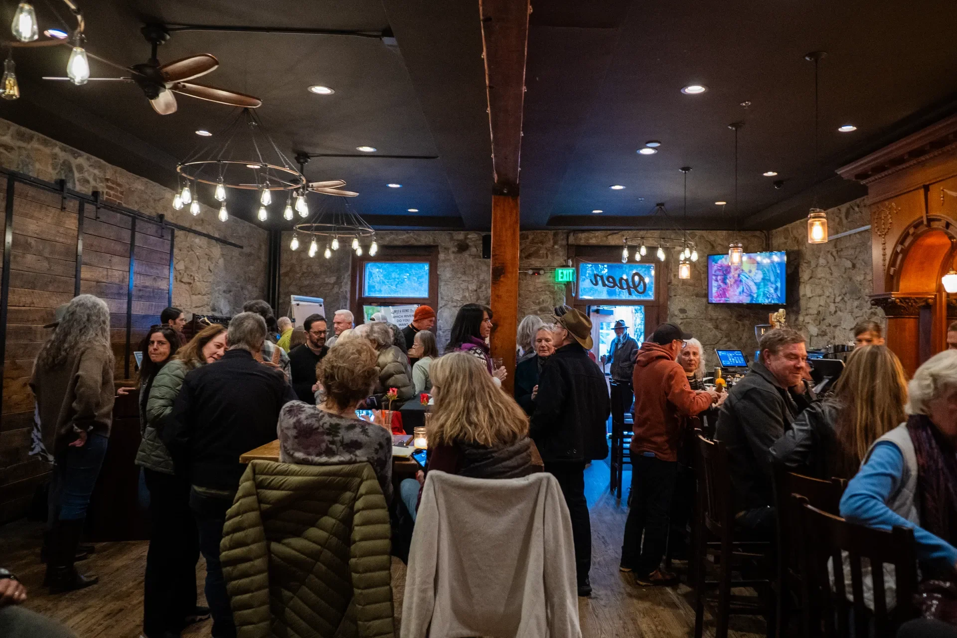 Crowded bar with people socializing, some seated at the bar and others standing. The interior features exposed brick walls, wooden accents, and warm lighting.