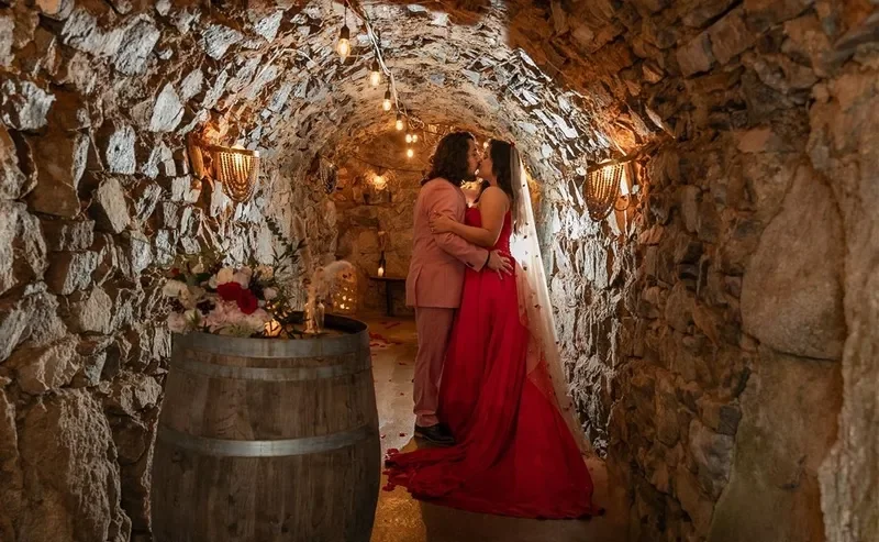 A couple sharing a kiss in a stone tunnel with warm lighting, decorated for a special occasion with flowers and a wine barrel.