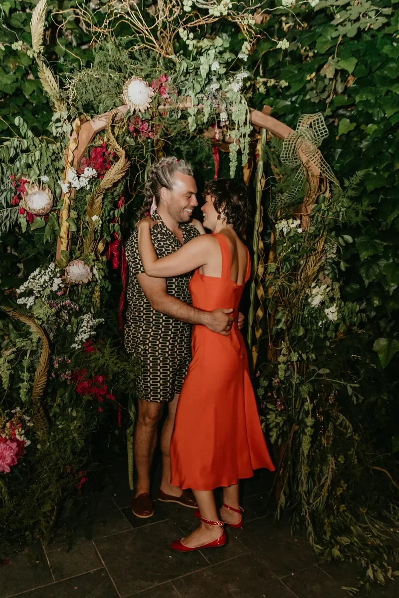 A couple embracing and smiling in front of a lush, decorated floral archway.
