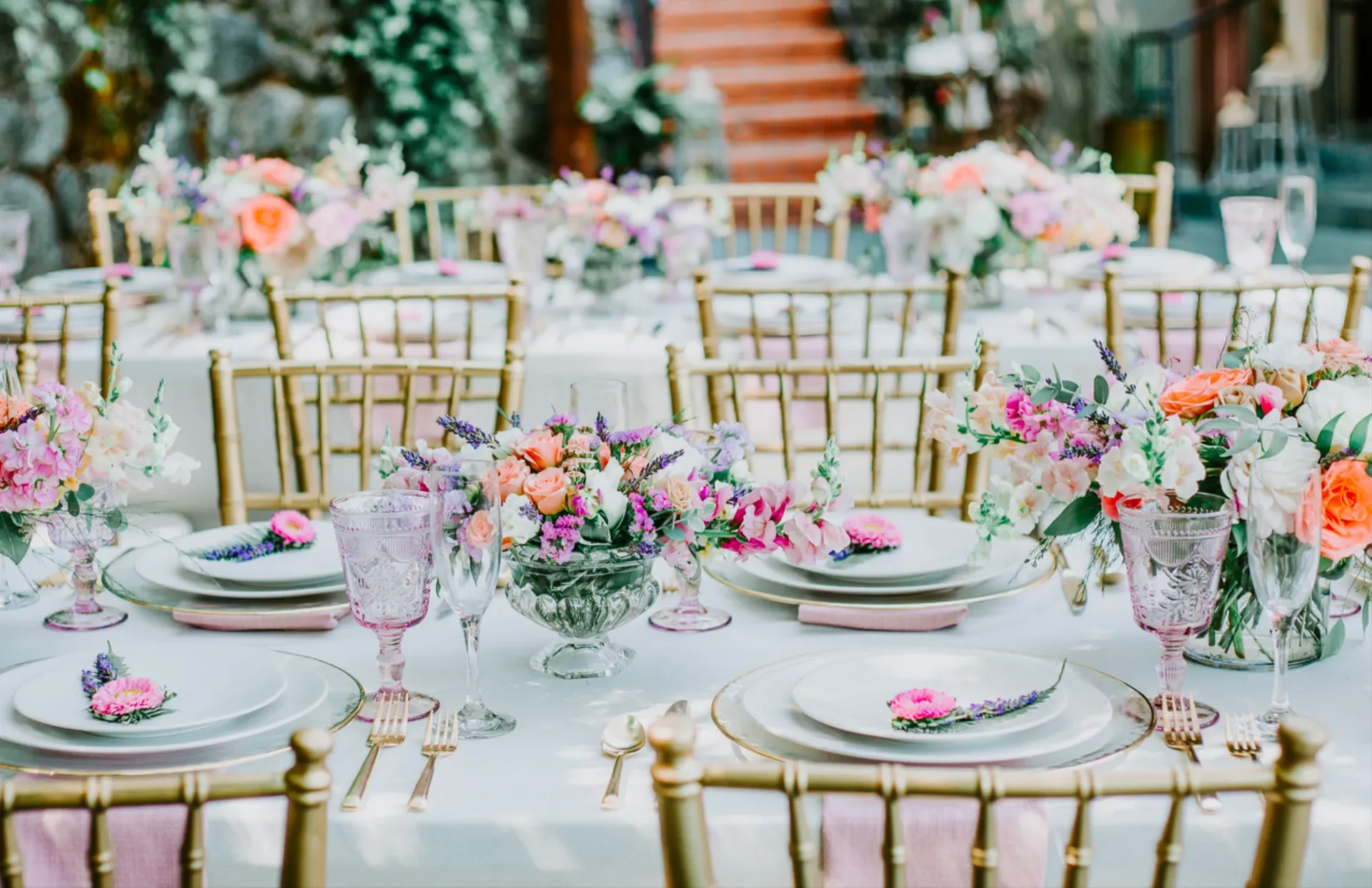 Elegant outdoor dining table set with floral centerpieces, pink and purple flowers, gold chairs, and pink napkins.