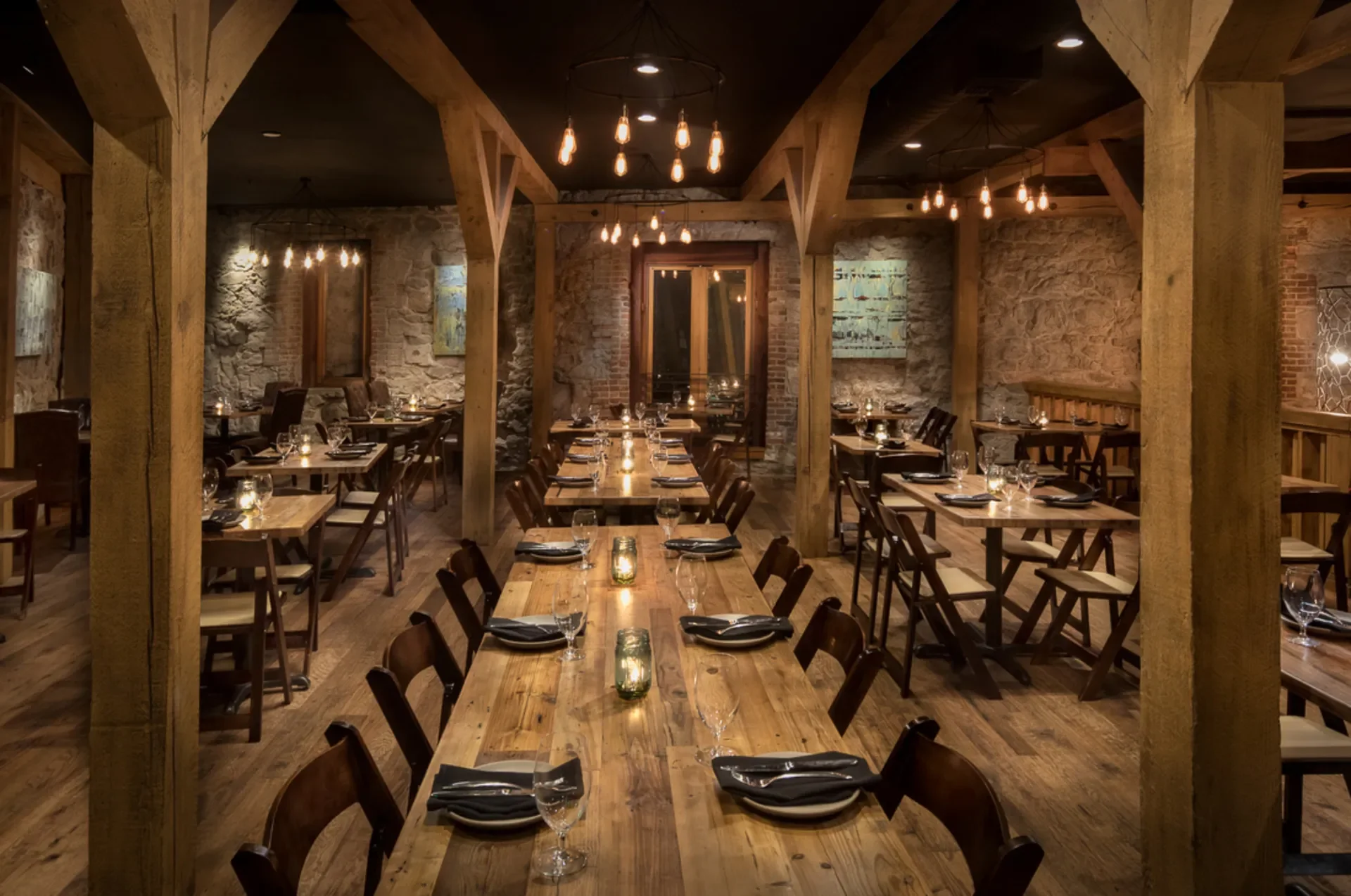 A cozy restaurant dining area with wooden tables, chairs, exposed brick and stone walls, modern hanging light fixtures, and candlelit place settings.