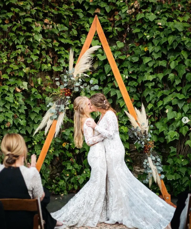 Two brides in white lace wedding dresses kiss in front of a wooden triangle arch decorated with greenery and dried flowers, with green foliage in the background, during a wedding ceremony.