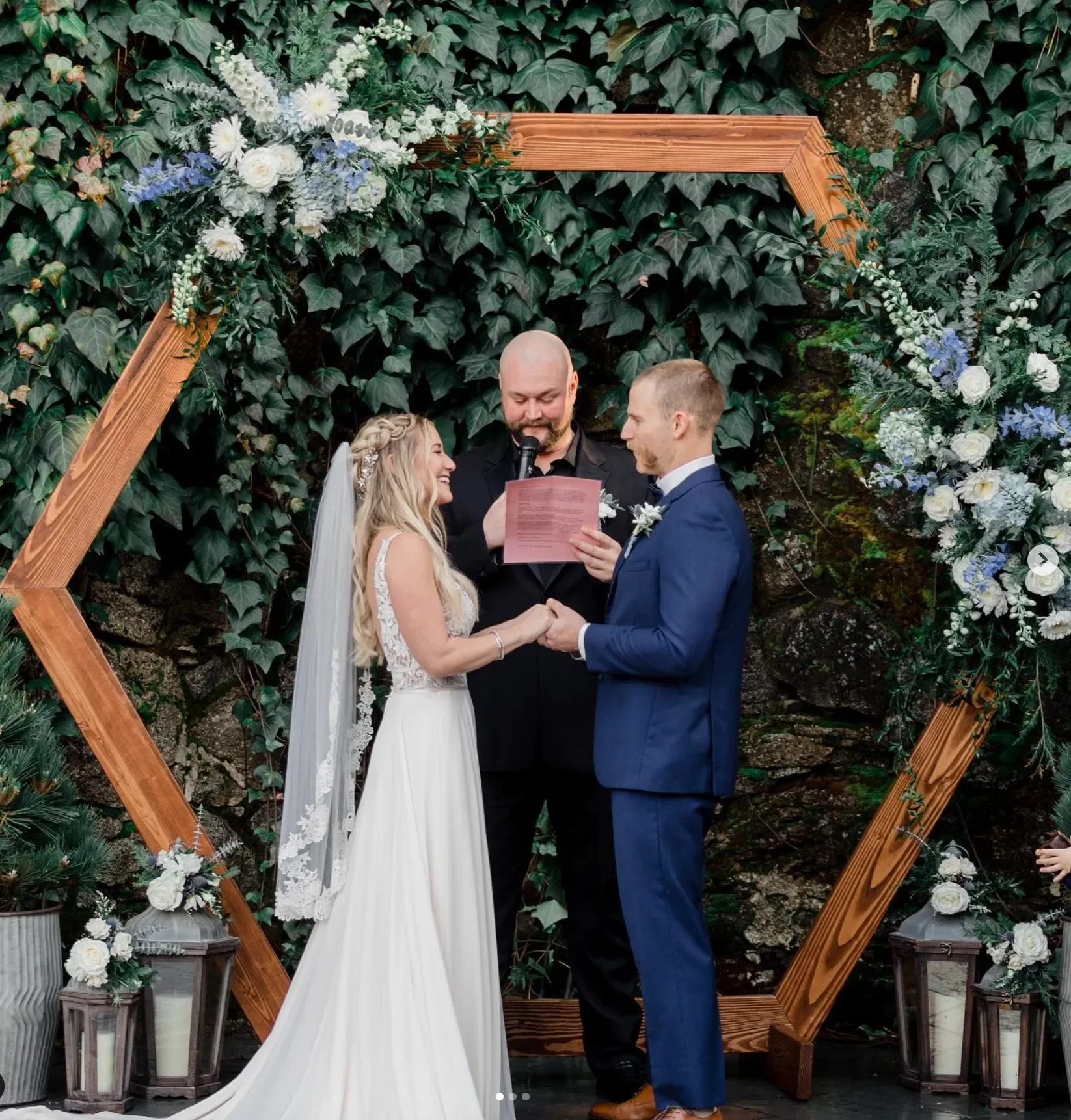 A couple getting married exchanging vows, with a officiant reading for them, in front of a decorative wooden hexagonal frame with flowers and greenery.