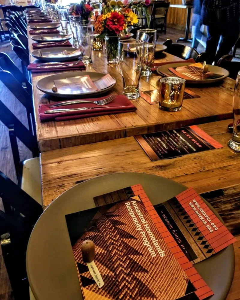 A long wooden table set for a meal with plates, wine glasses, water glasses, gold-colored candle holders, and colorful floral centerpieces. Menus are placed at each setting. The table has napkins and silverware arranged neatly.
