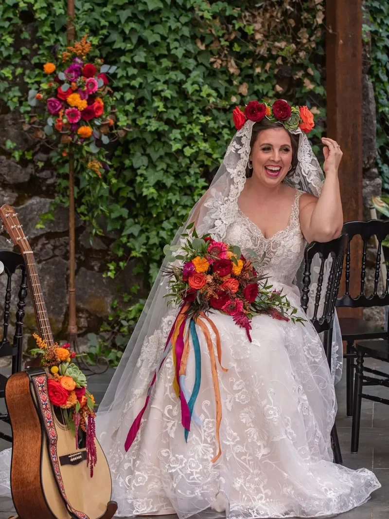 A bride in a white lace wedding dress and veil, wearing a floral crown, sitting and smiling, holding a colorful bouquet of flowers with ribbons, in a garden setting with ivy-covered wall and chairs.