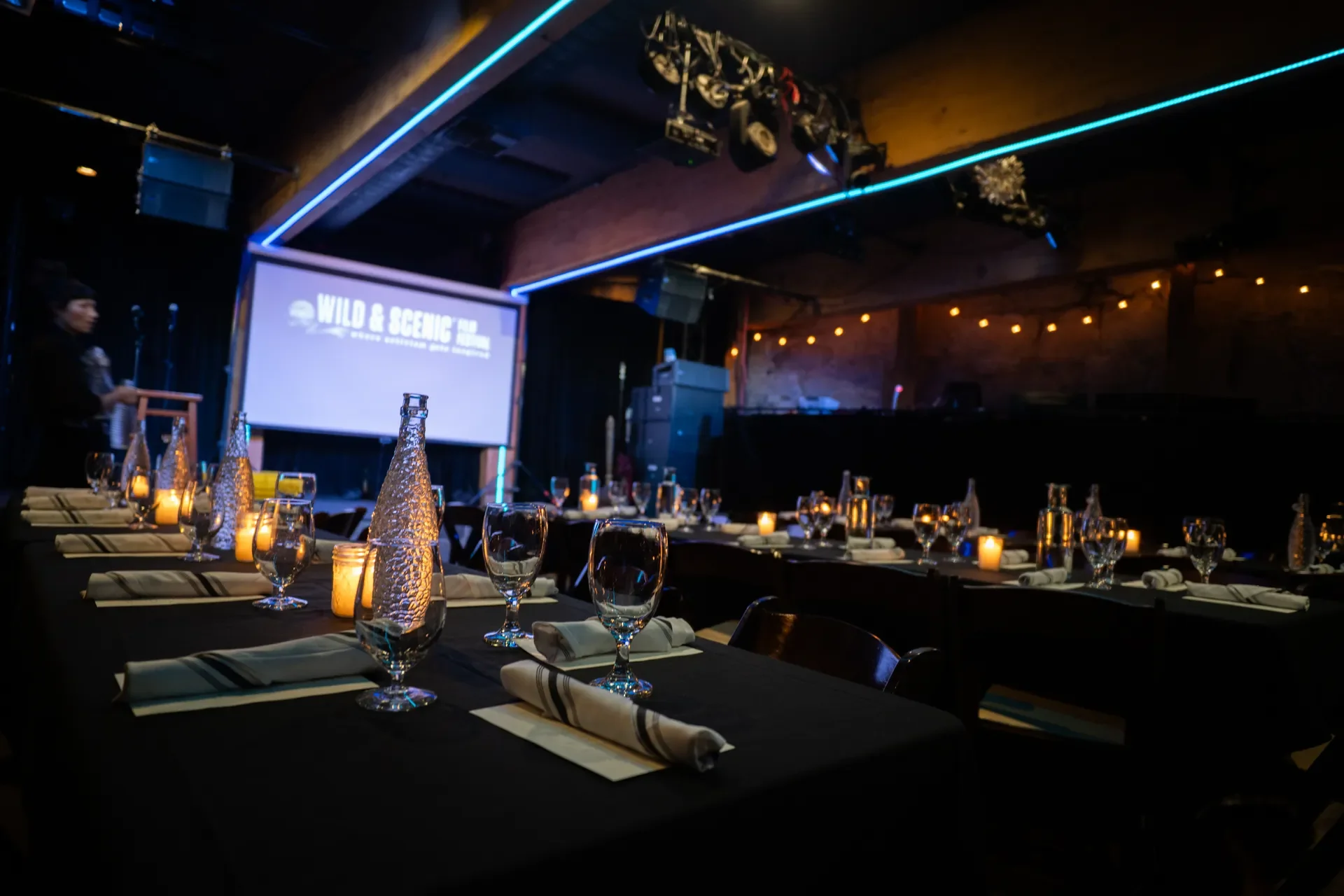 Dimly lit event space with tables set for dining, decorated with candles, glassware, and napkins, and a large screen displaying 'Wild & Scenic' in the background.
