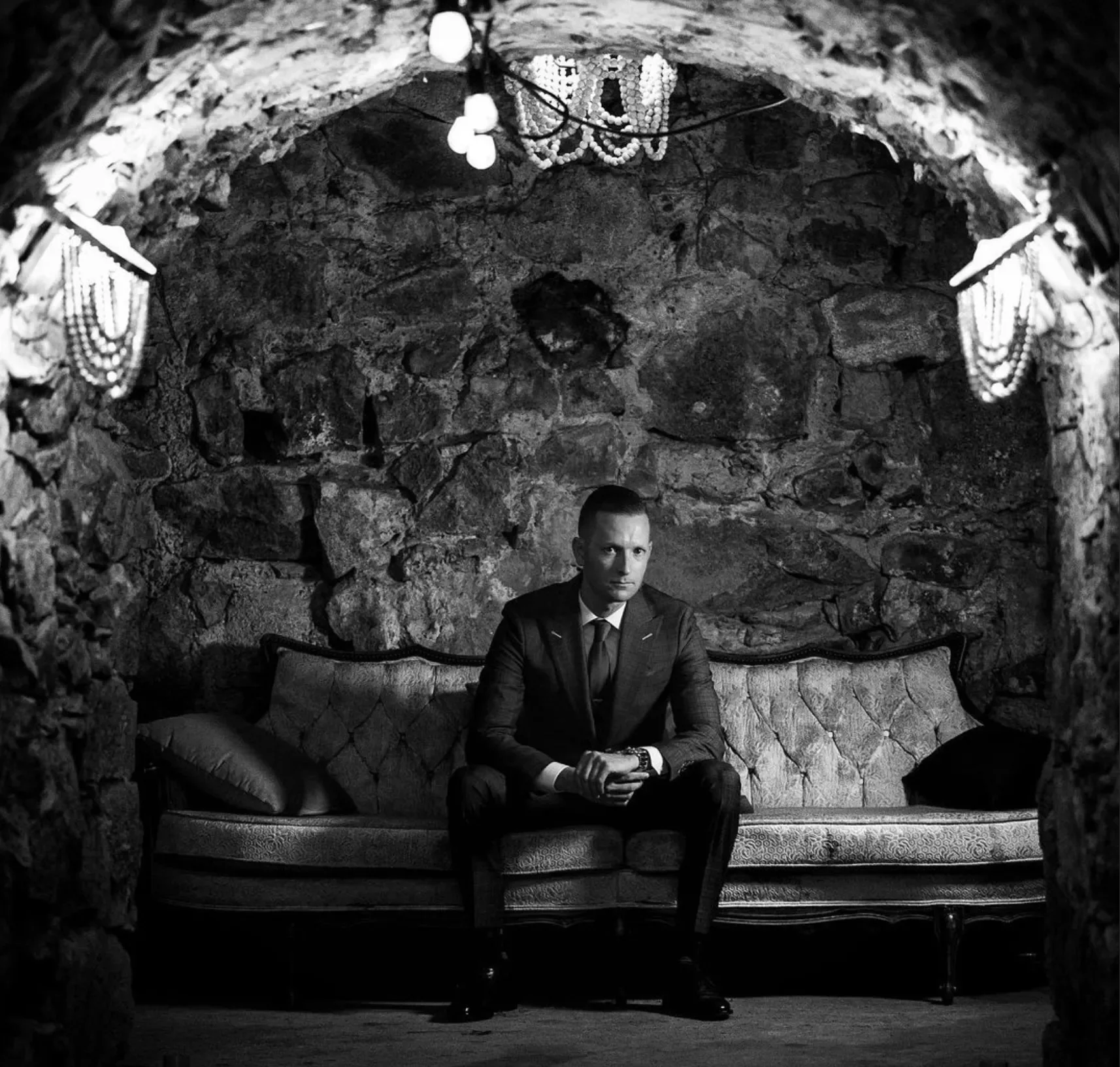 A man in a suit sitting on an ornate vintage sofa in a stone-walled room with hanging chandeliers and jewelry decoration.