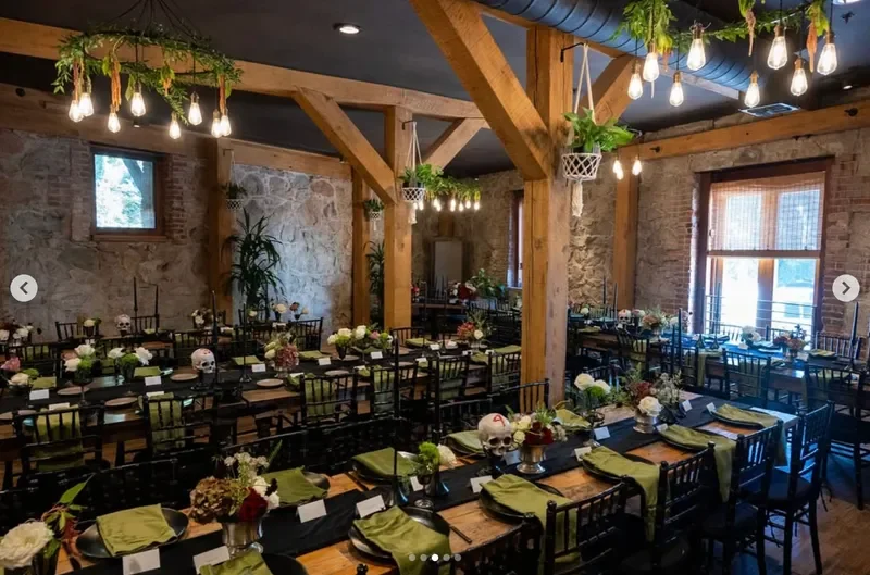 Event venue with long wooden tables decorated with floral arrangements, skull accents, and green napkins, in a rustic brick and wood interior with string lights overhead.