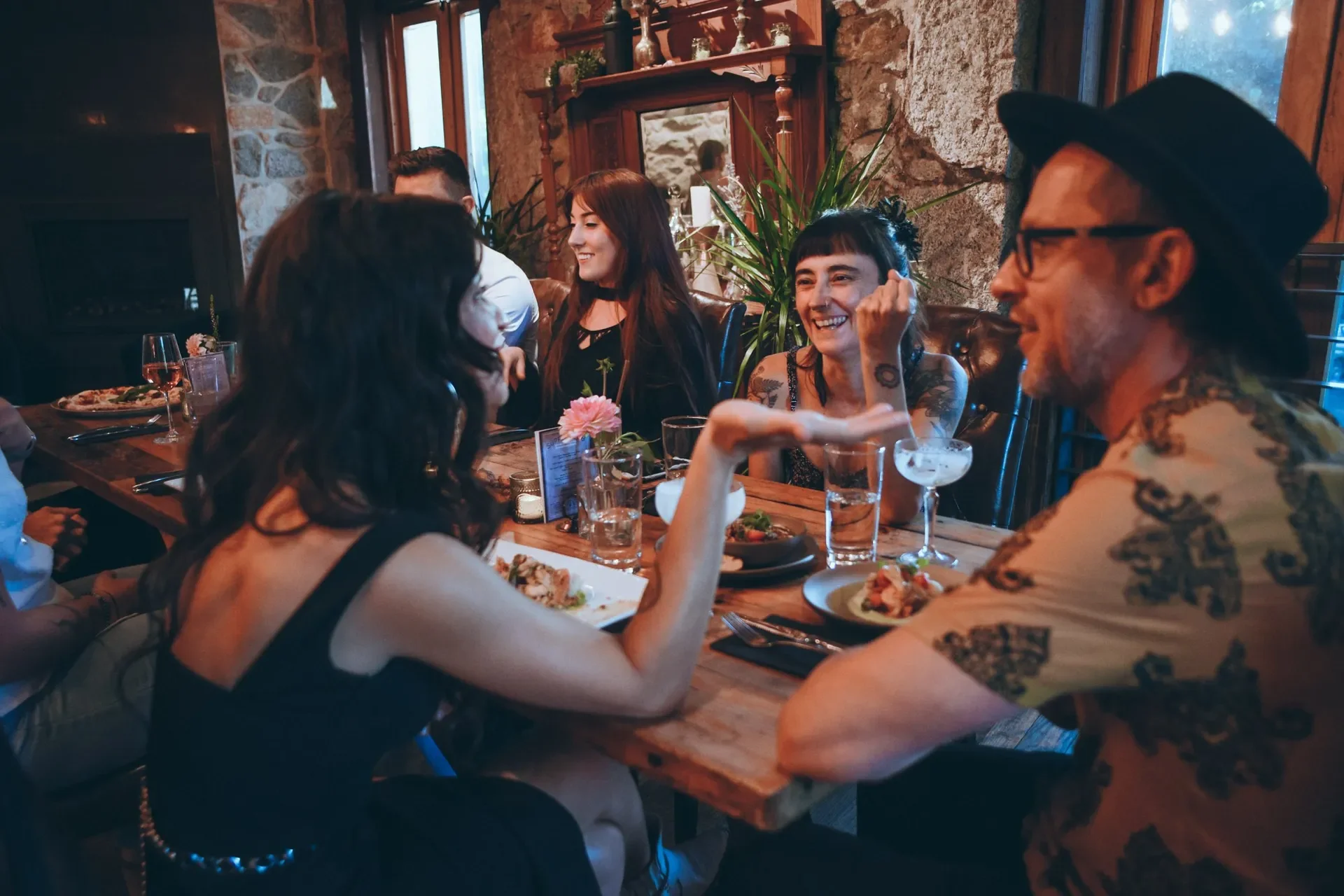 A group of friends sitting at a dining table, enjoying food and drinks, in a cozy restaurant with stone walls and wooden accents. They are smiling and engaging in conversation, with some people leaning on their hands and one man wearing a hat.