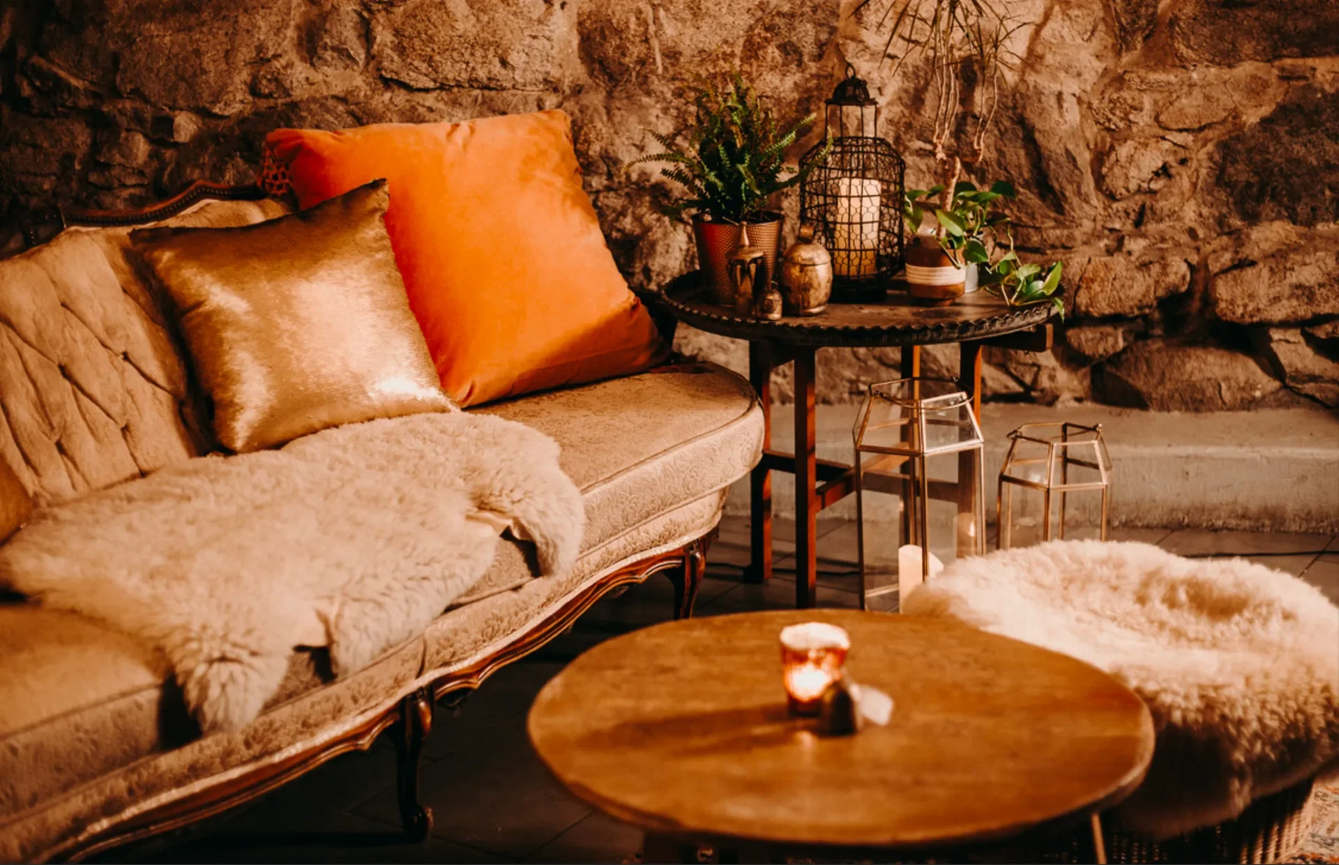 Cozy living room with vintage sofa adorned with orange and metallic pillows, furry blanket, and sheepskin rug, a round wooden coffee table with a lit candle, and a side table with plants, lanterns, and decorative items against a stone wall.
