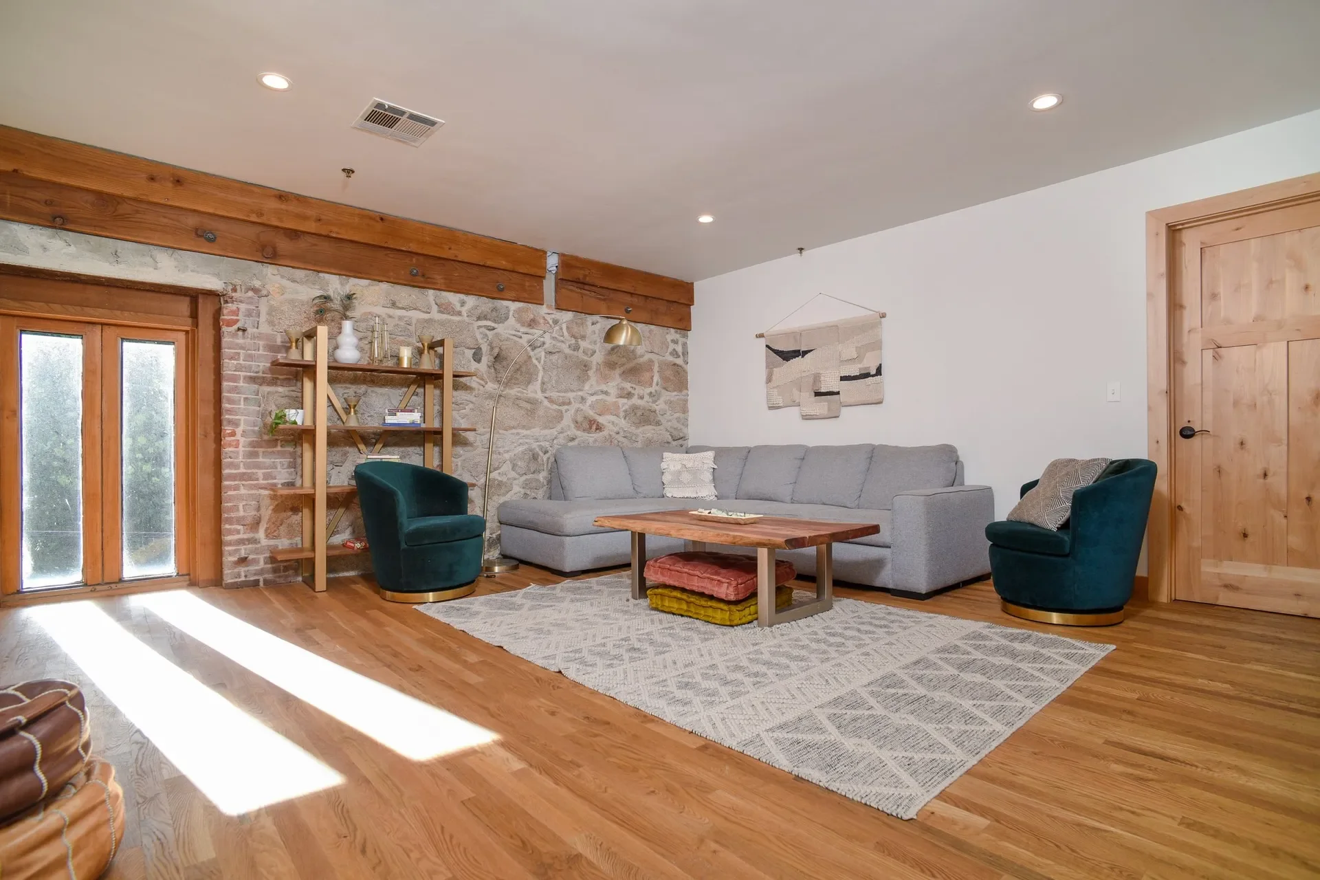 Living room with wooden floors, stone wall, gray sofa, two dark green armchairs, a wooden coffee table with stacked cushions underneath, a tall floor lamp, a wooden bookshelf with decorative items, and a wall hanging art piece.