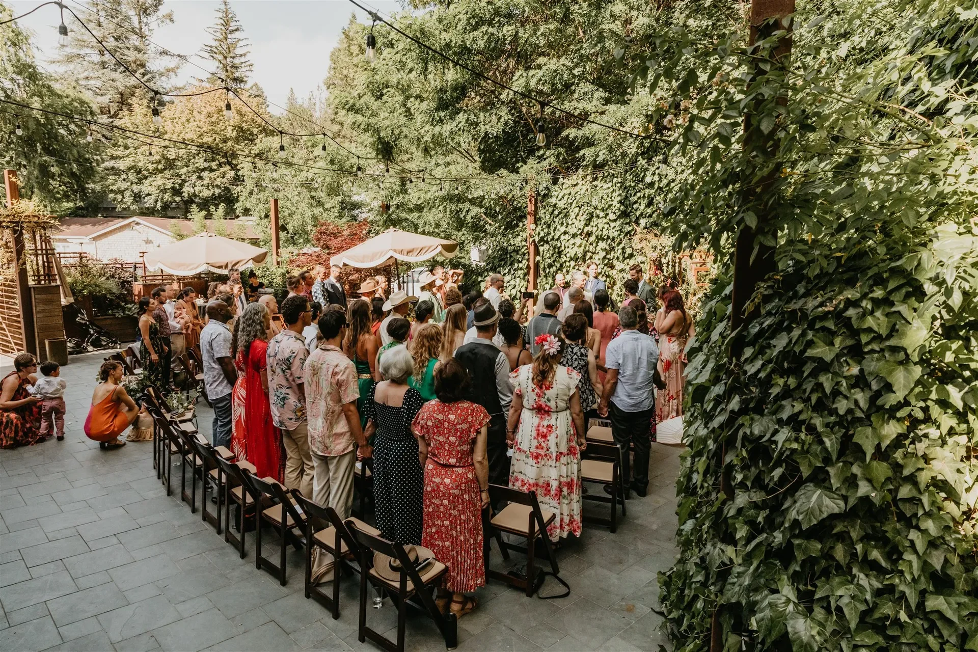 Group of people dressed in colorful attire attending an outdoor gathering or celebration in a garden with trees, ivy, and string lights.
