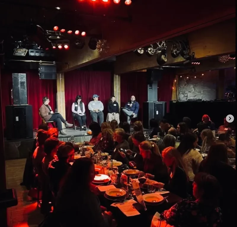 A panel of five people sitting on stage with a red curtain backdrop, microphones, and audio equipment, addressing an audience in a dimly lit venue with red stage lighting. The audience is seated at tables with plates, glasses, and utensils.