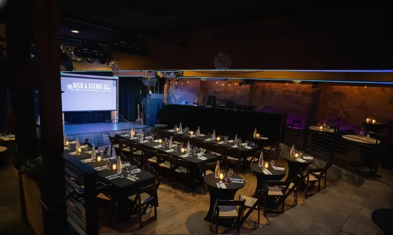 Empty banquet-style dining area with candlelit tables, a large screen displaying 'Wild and Scenic All...' in a music venue or event space.