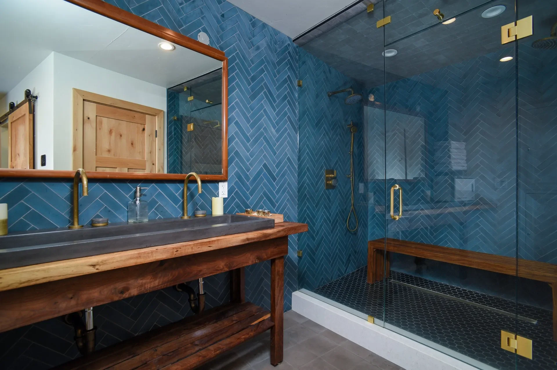 Modern bathroom with blue herringbone tile walls, a clear glass shower enclosure with brass handles and fixtures, a wooden vanity with a dark countertop, a large mirror with a wooden frame, and a wooden door.