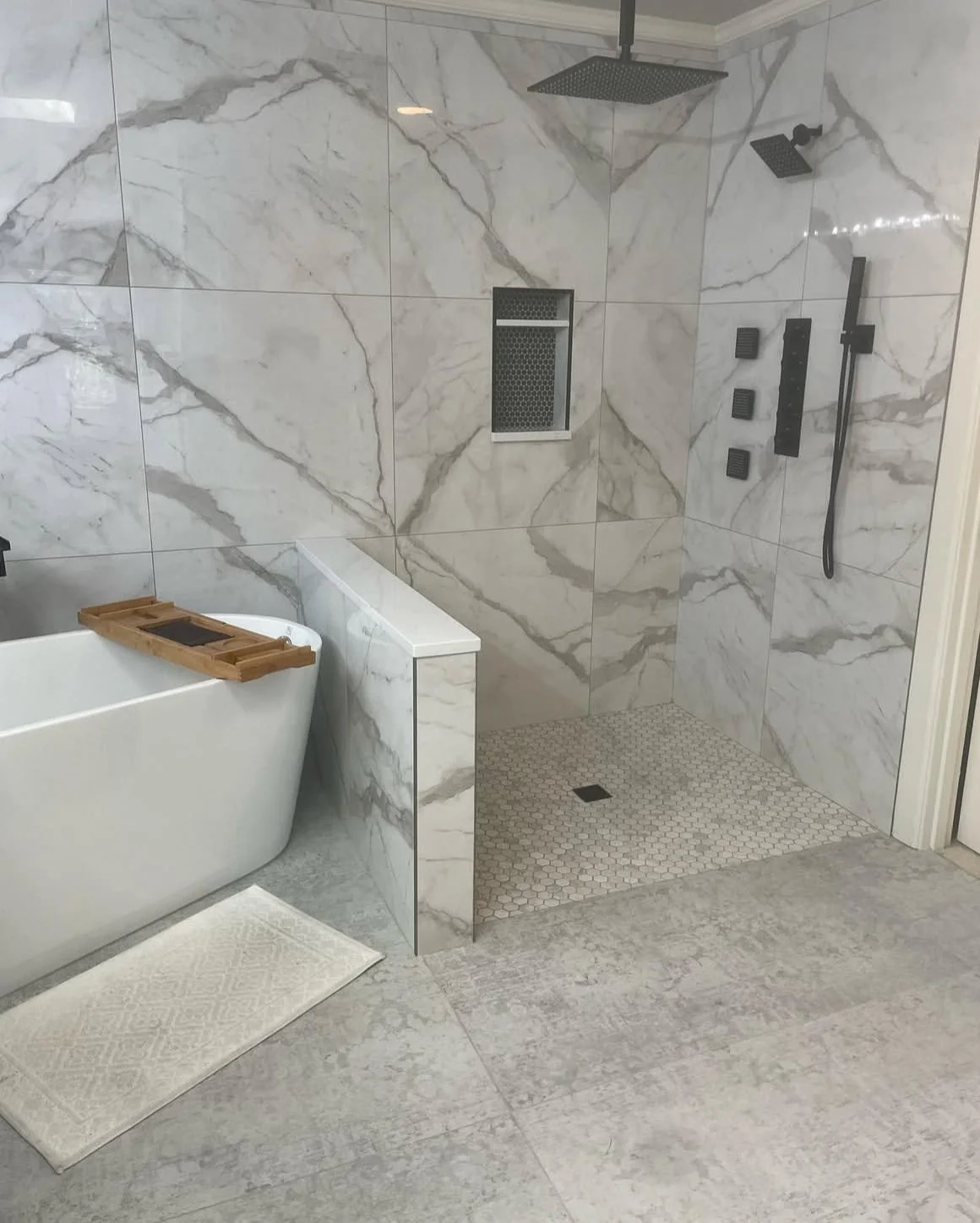 Modern bathroom with marble tile shower area, black shower fixtures, and a separate bathtub with a wooden tray on top, beige patterned rug, and a textured concrete floor.