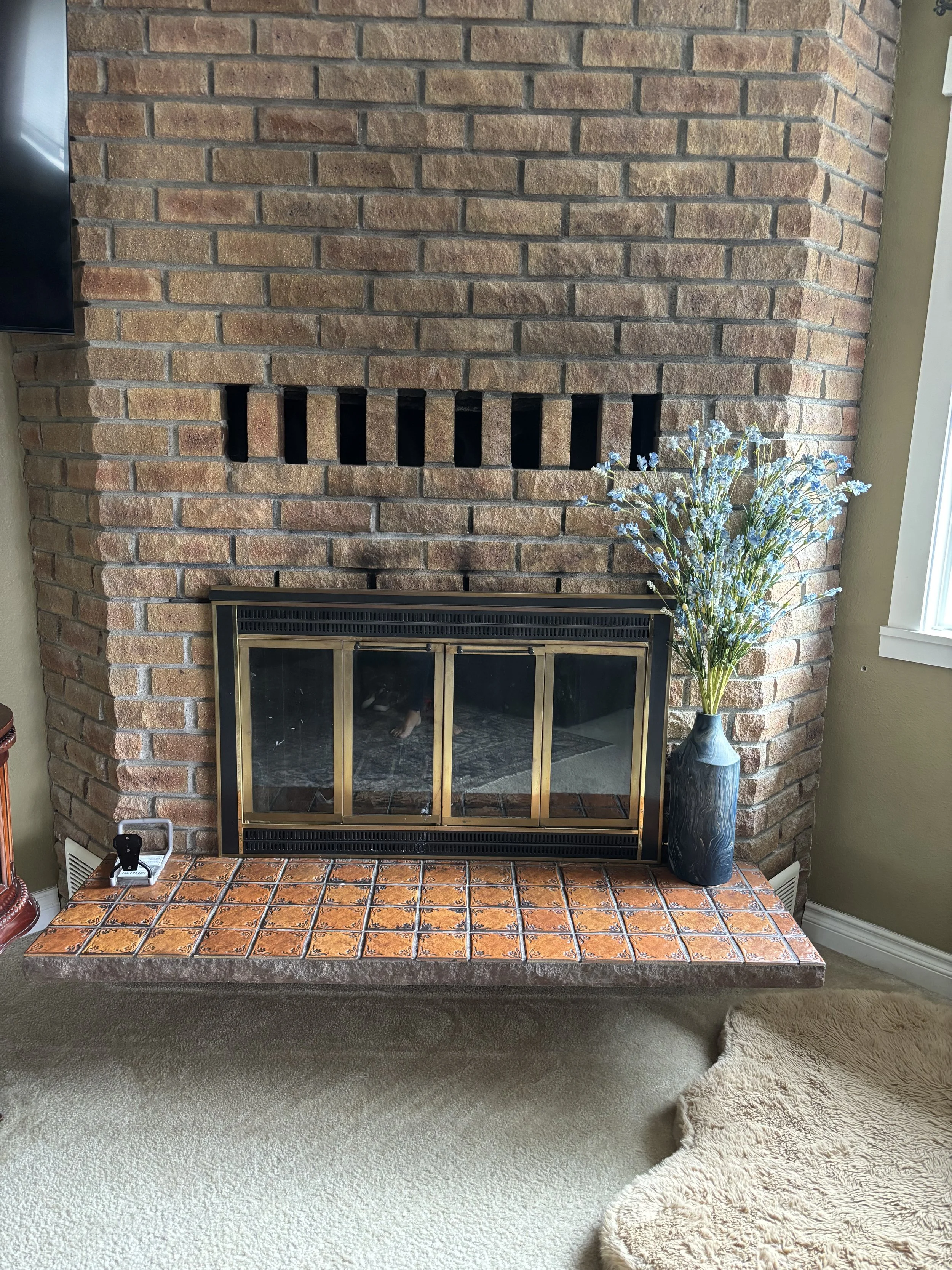 Brick fireplace with a glass door, a tall blue vase with artificial flowers, and a small black object on a ledge with orange tiles. Carpeted floor with a partial beige rug, nearby window, and a wall vent.