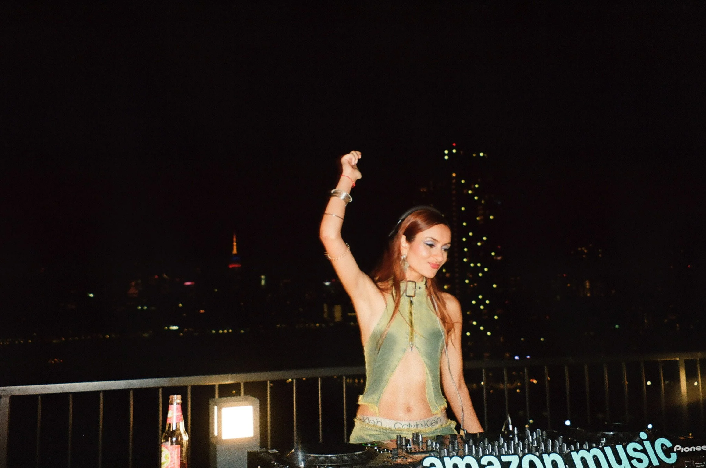 Vani Dewan DJing and dancing with one arm raised at night, city skyline in background, DJ equipment in front, illuminated by outdoor lights.