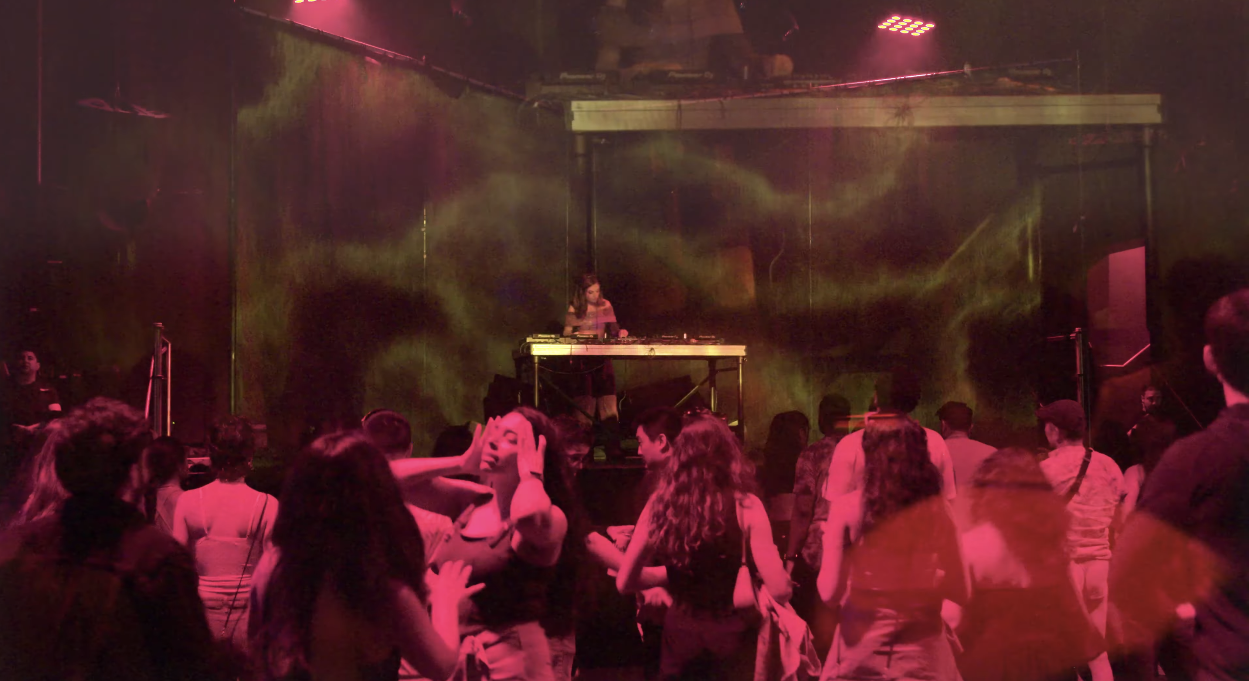 People dancing at a nightclub with a DJ on stage, colorful lighting, and smoke effects.