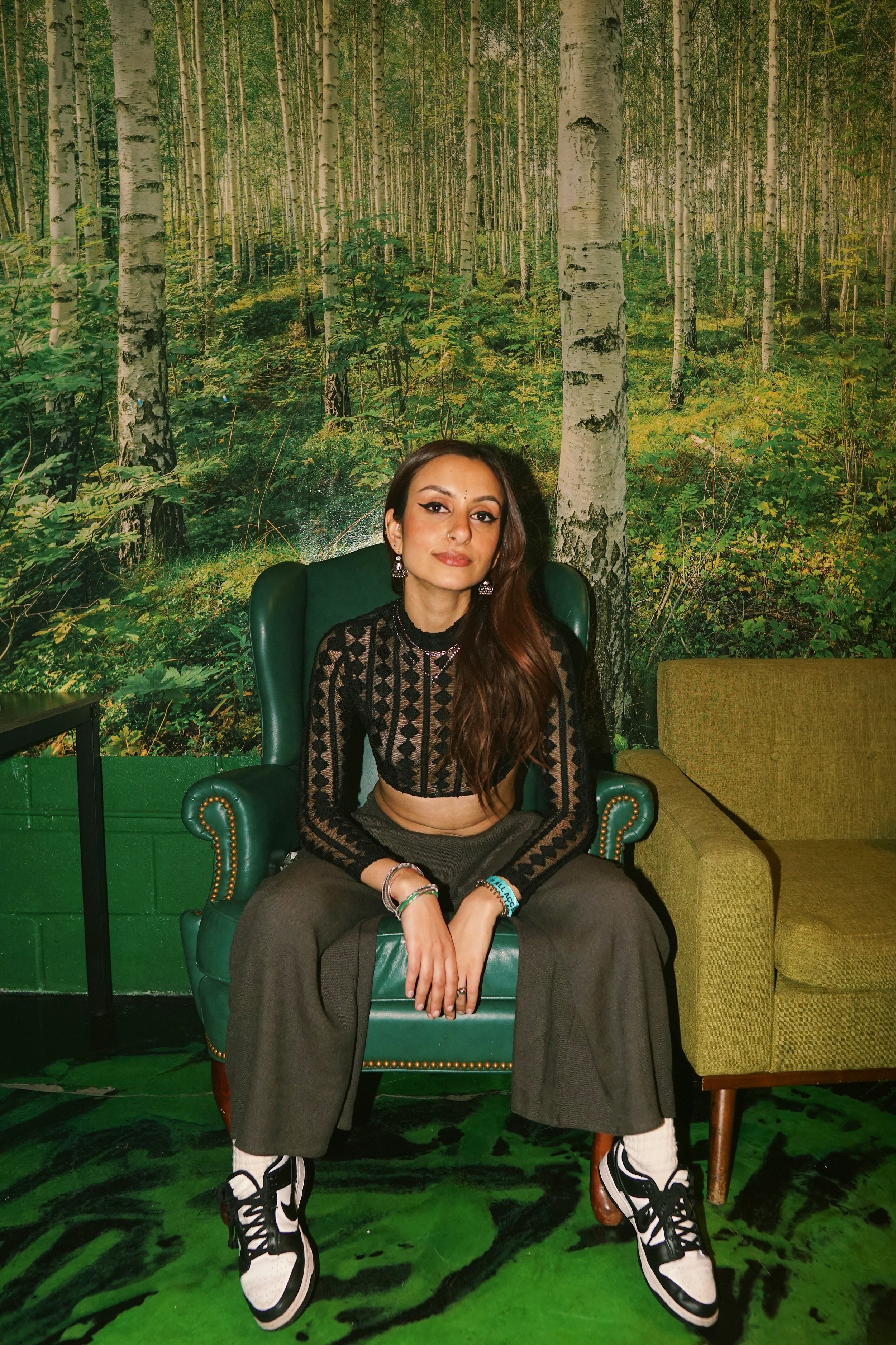 Vani sits in a dark green chair against a forest background.