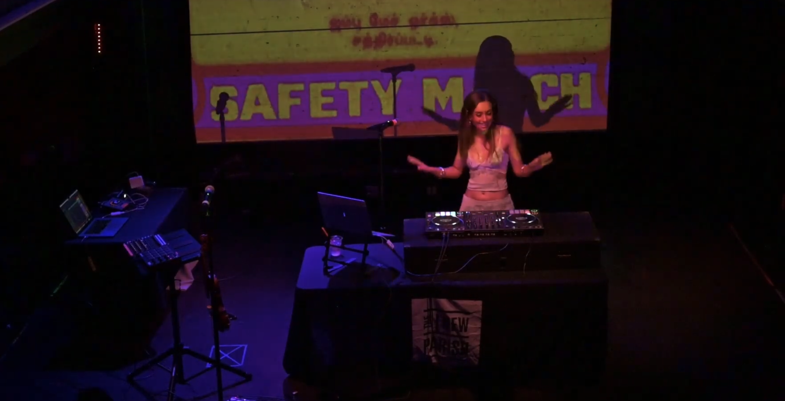 Vani Dewan performing on stage at the New Parish in Oakland with audio equipment and a laptop under purple and red lighting. A large screen behind displays a yellow safety match advertisement.