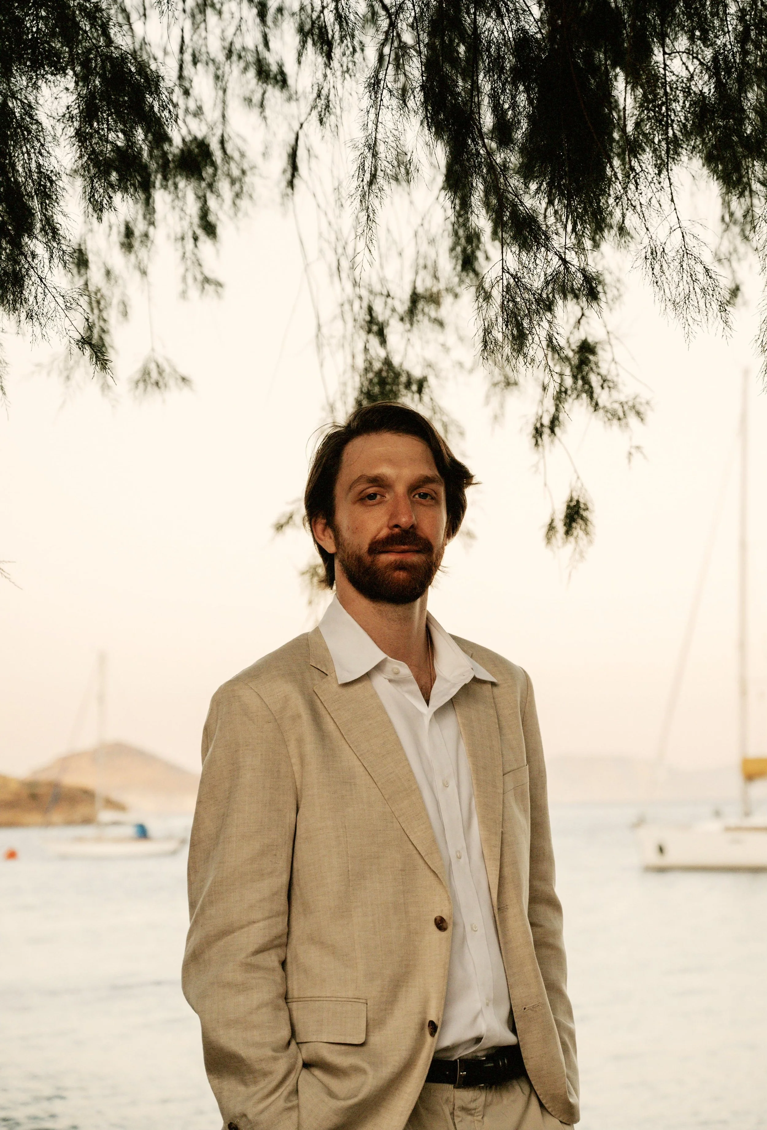 A man with dark hair and a beard wearing a beige suit standing near water with boats in the background and tree branches overhead.