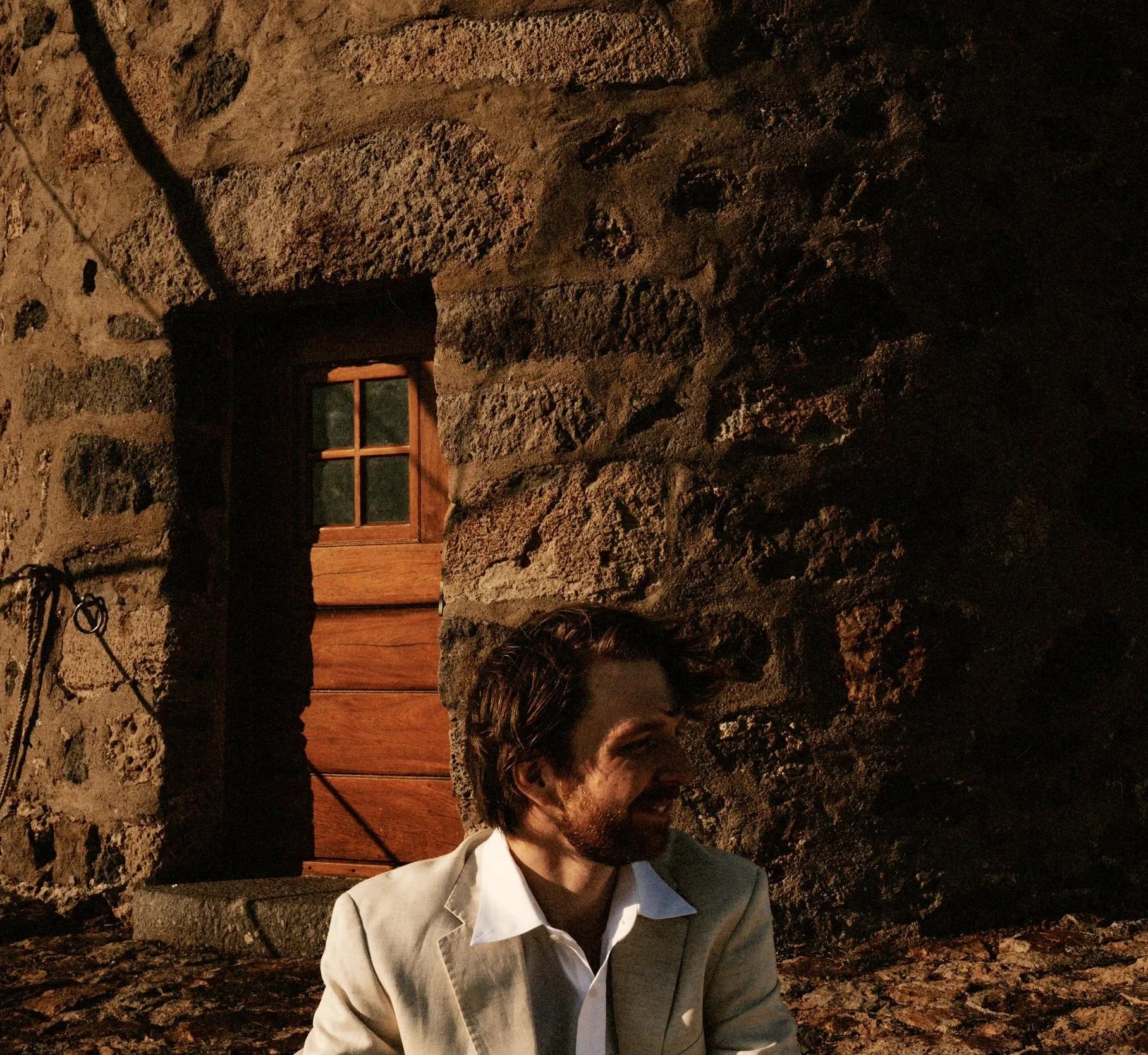 A man in a beige suit with a white shirt sits outdoors near a stone wall and wooden door, smiling and looking to his right during sunset.